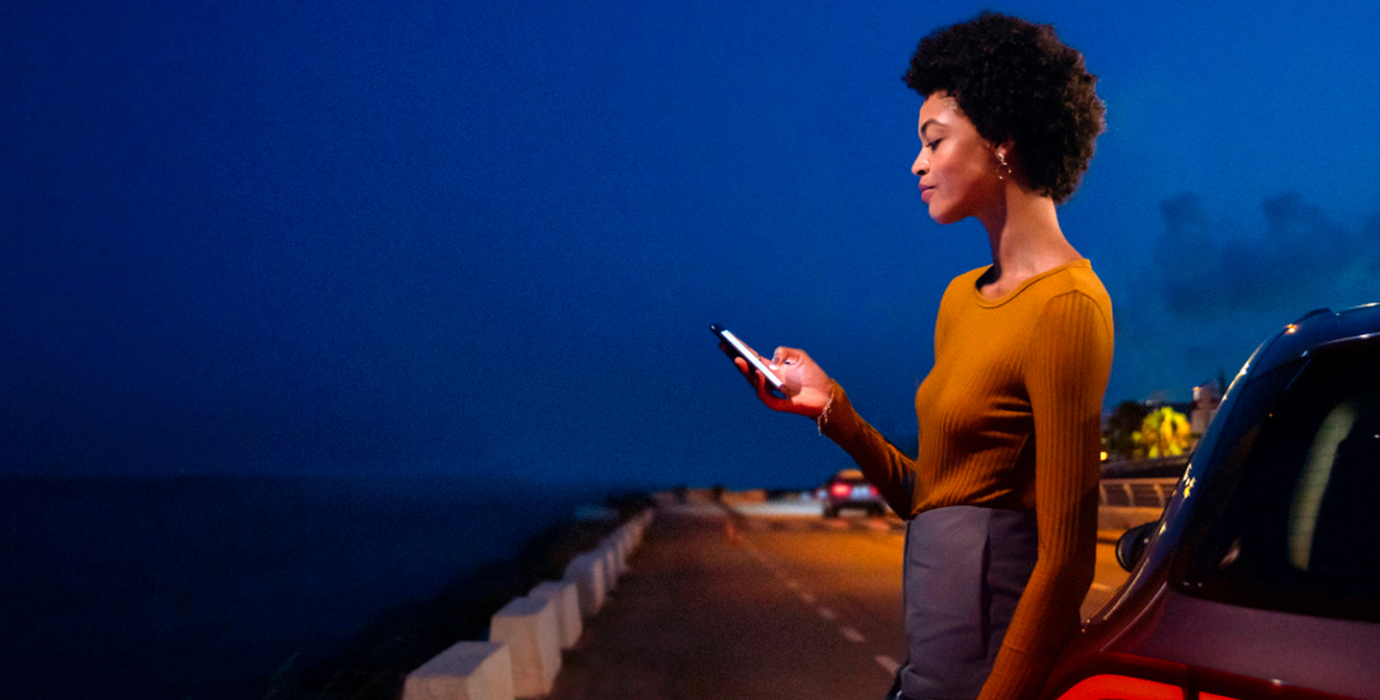 Neuwagen kaufen - so geht's! | Mercedes-Benz Eine Frau in gelbem Outfit schaut auf ihr Handy und steht dabei an einem Straßenrand.