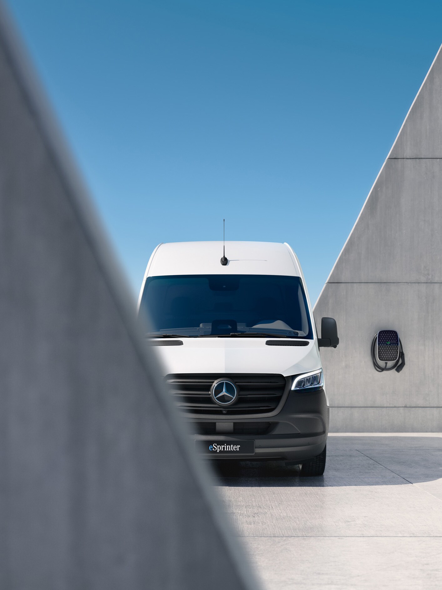 Ein vollelektrischer eSprinter in Frontansicht vor und hinter einer grauen Mauer. Im Hintergrund eine Wallbox.