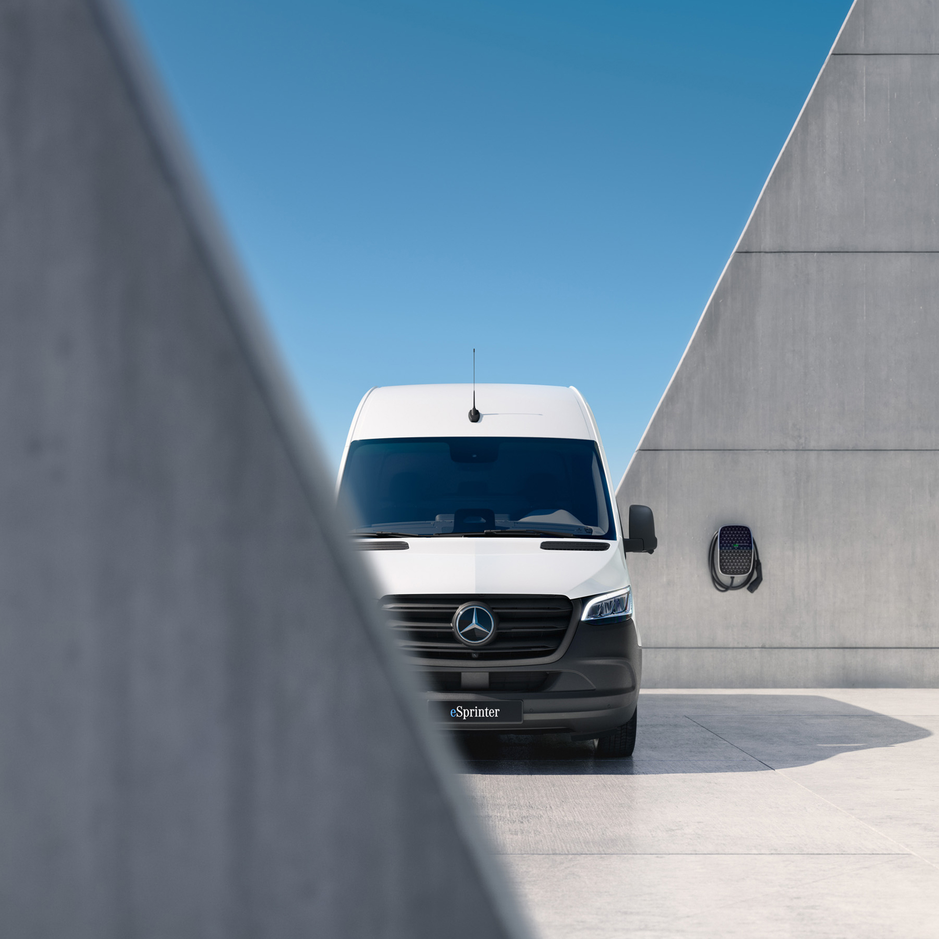 Mercedes-Benz | eSprinter Kastenwagen Ein vollelektrischer eSprinter in Frontansicht vor und hinter einer grauen Mauer. Im Hintergrund eine Wallbox.