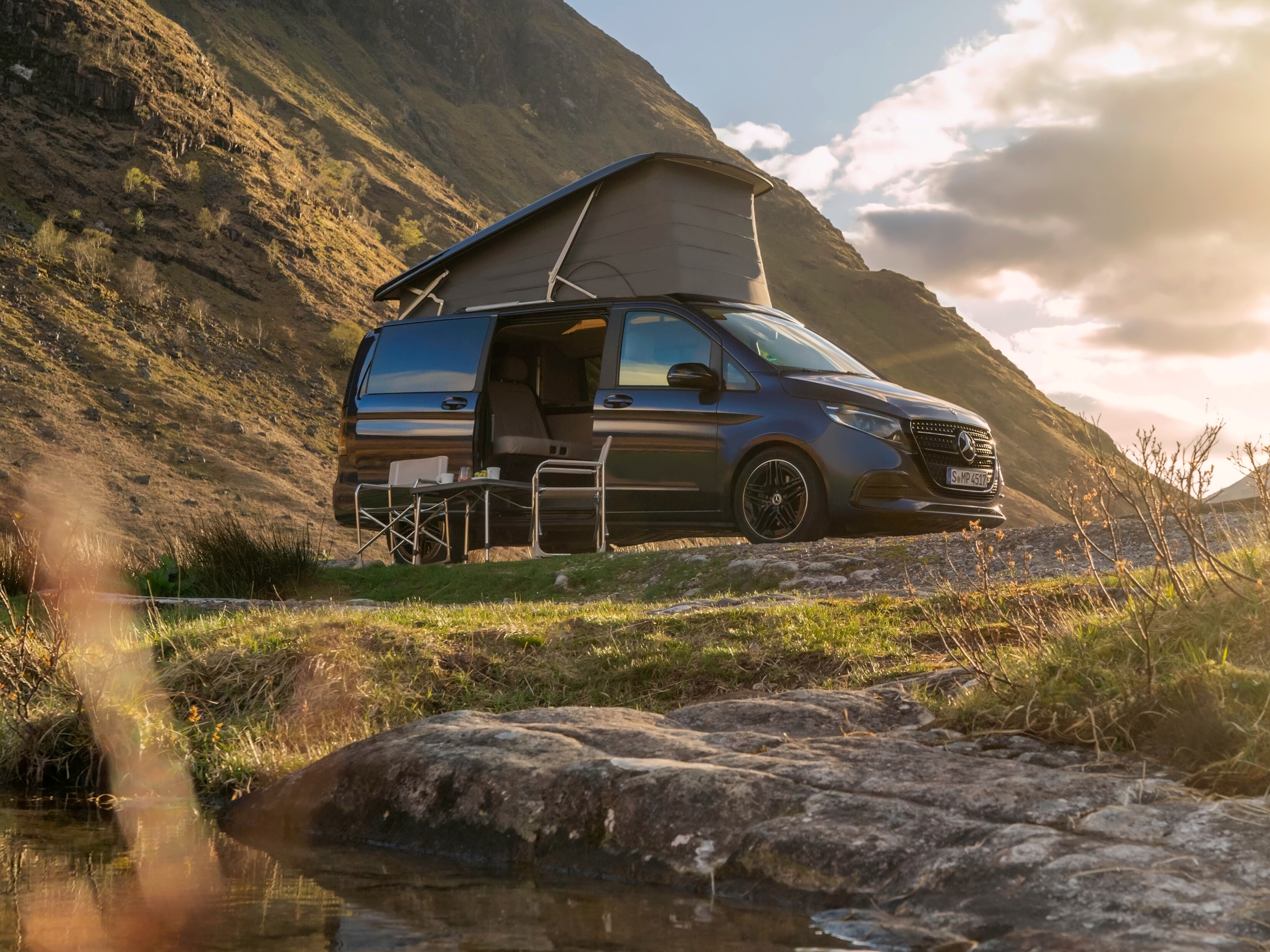 Ein Mercedes-Benz Camper steht mit offenem Dach in der Natur. Vor der geöffneten Schiebetür stehen ein Campingtisch und zwei Stühle. 