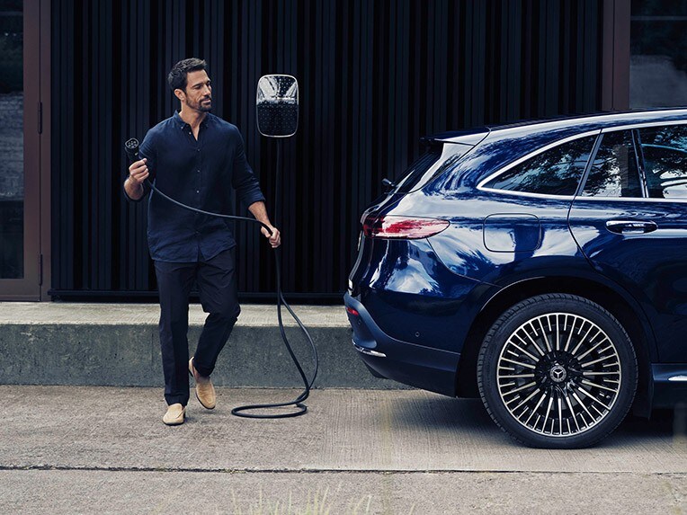 Mann geht hinter blauem Mercedes Benz EQC mit Fahrzeugladekabel