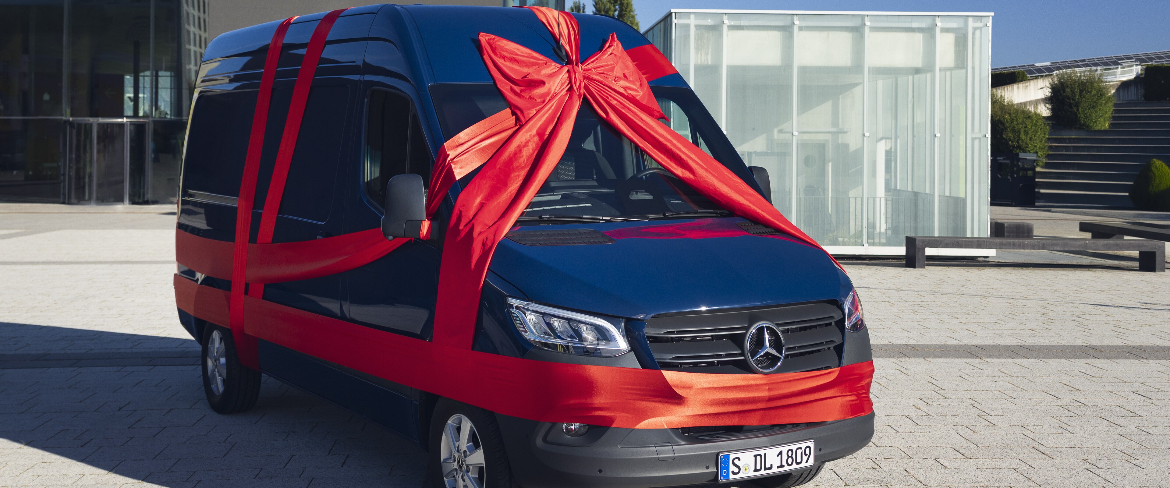 Blauer Sprinter Kastenwagen mit einer roten Schleife umhüllt. Parkend vor einem Gebäude.