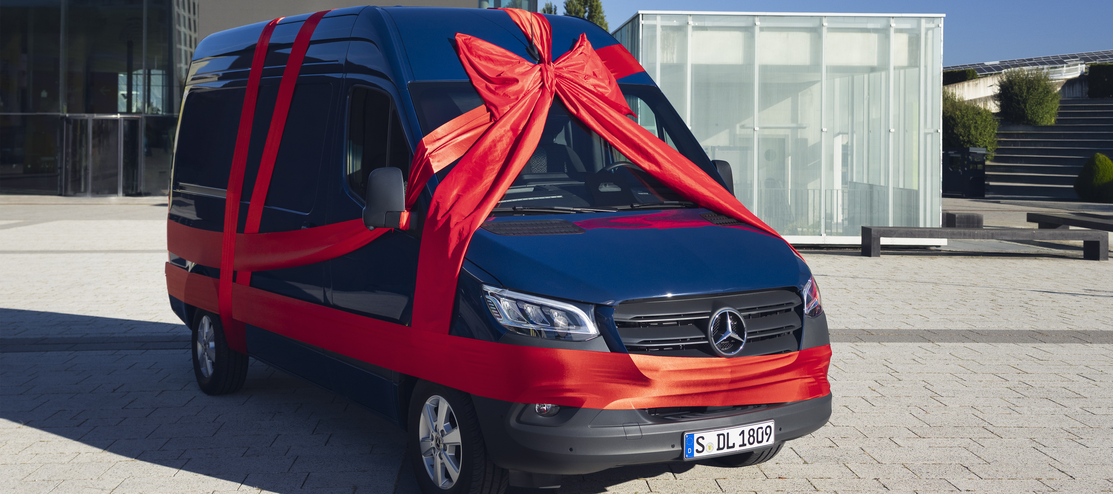 Happy Holidays | Mercedes-Benz Vans Blauer Sprinter Kastenwagen mit einer roten Schleife umhüllt. Parkend vor einem Gebäude.