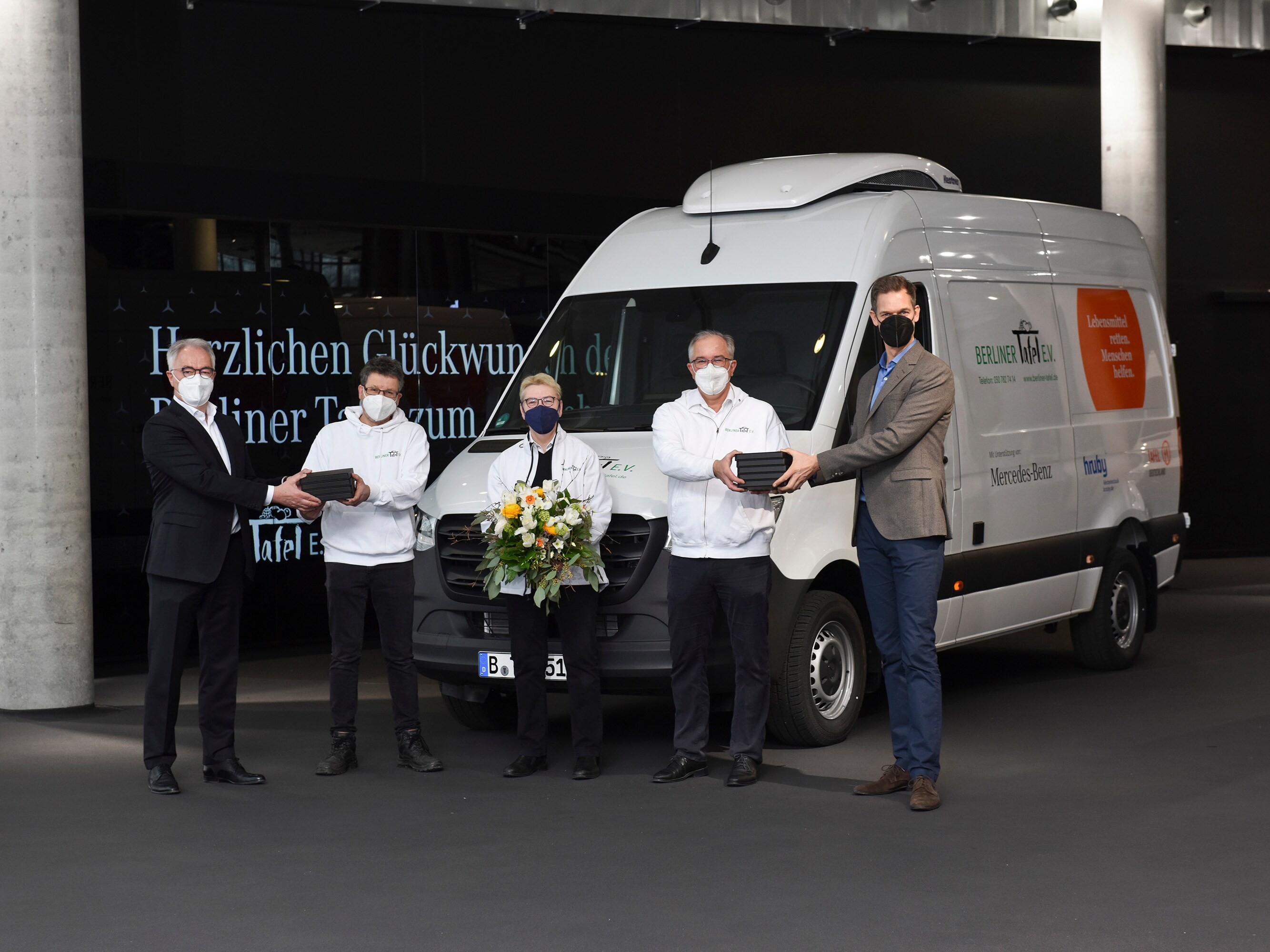 Fünf Personen stehen vor einem Mercedes-Benz Sprinter feierlich zu einer offiziellen Übergabe. Eine Person hält einen Blumenstrauß in die Hände.