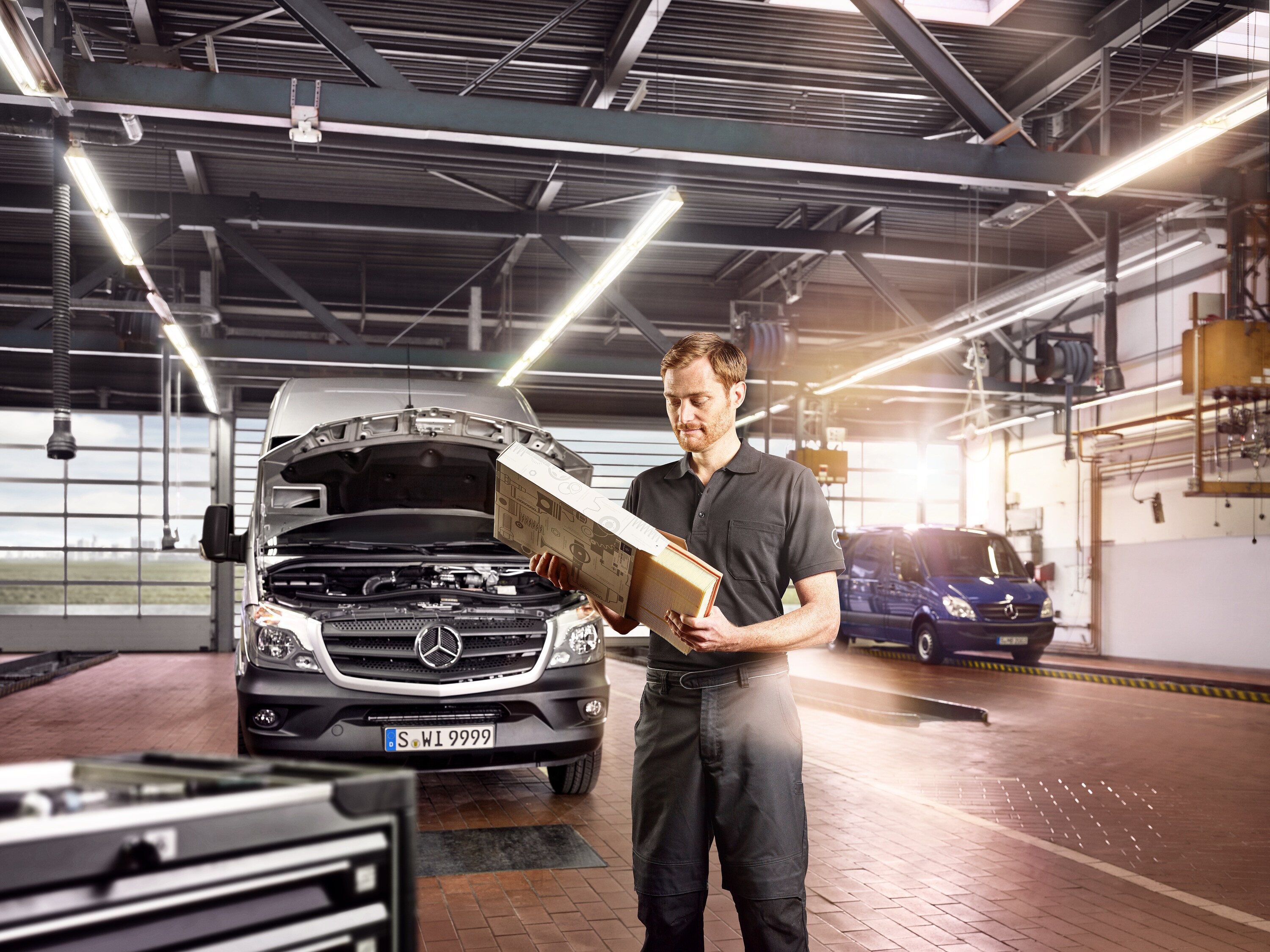 Ein Mercedes-Benz Servicemitarbeiter hält ein Paket in den Händen. Ein Transporter steht im Hintergrund mit geöffneter Motorhaube.