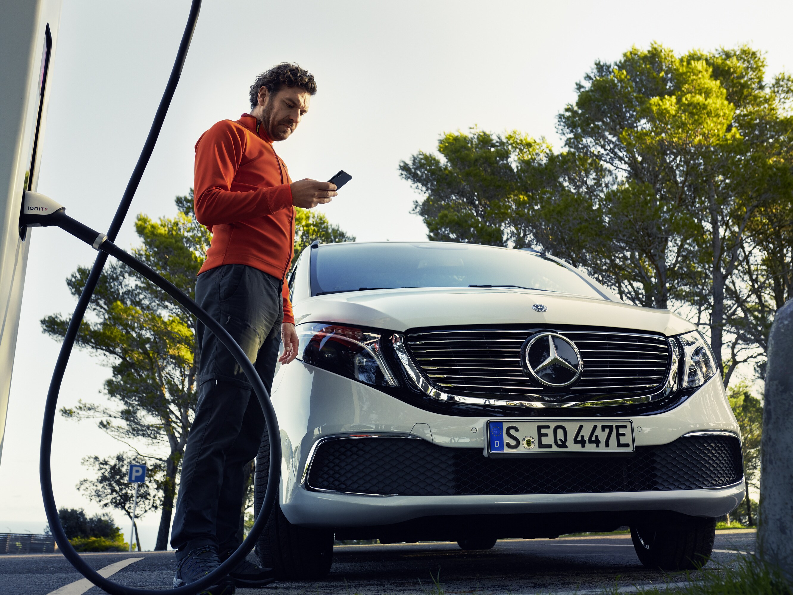 Eine Person schaut auf ihr Smartphone während ein vollelektrischer Mercedes-Benz Van an der Ladesäule steht. 