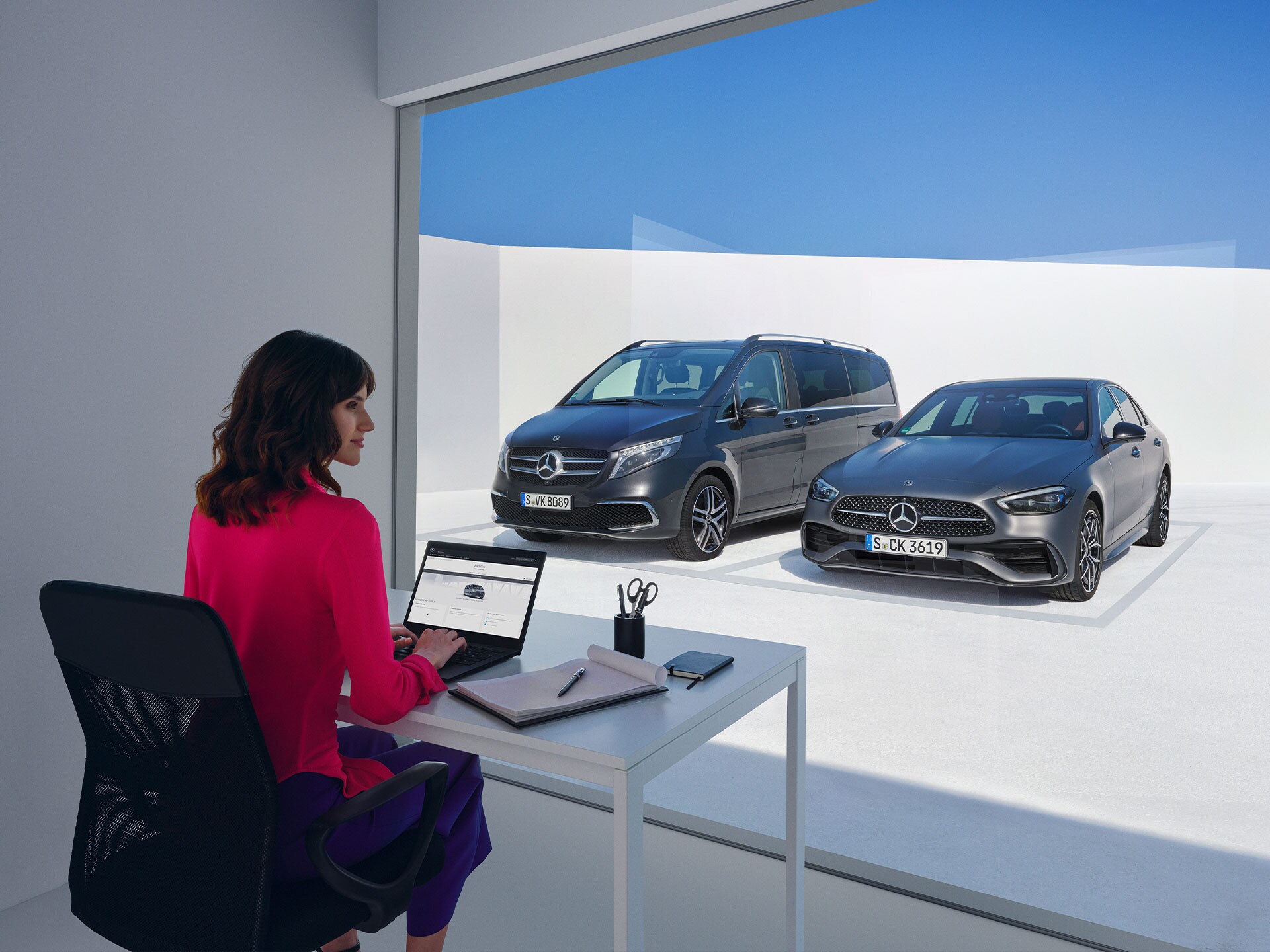 Eine Frau sitzt vor einer Mercedes-Benz V-Klasse und C-Klasse und schaut auf einen Laptop.