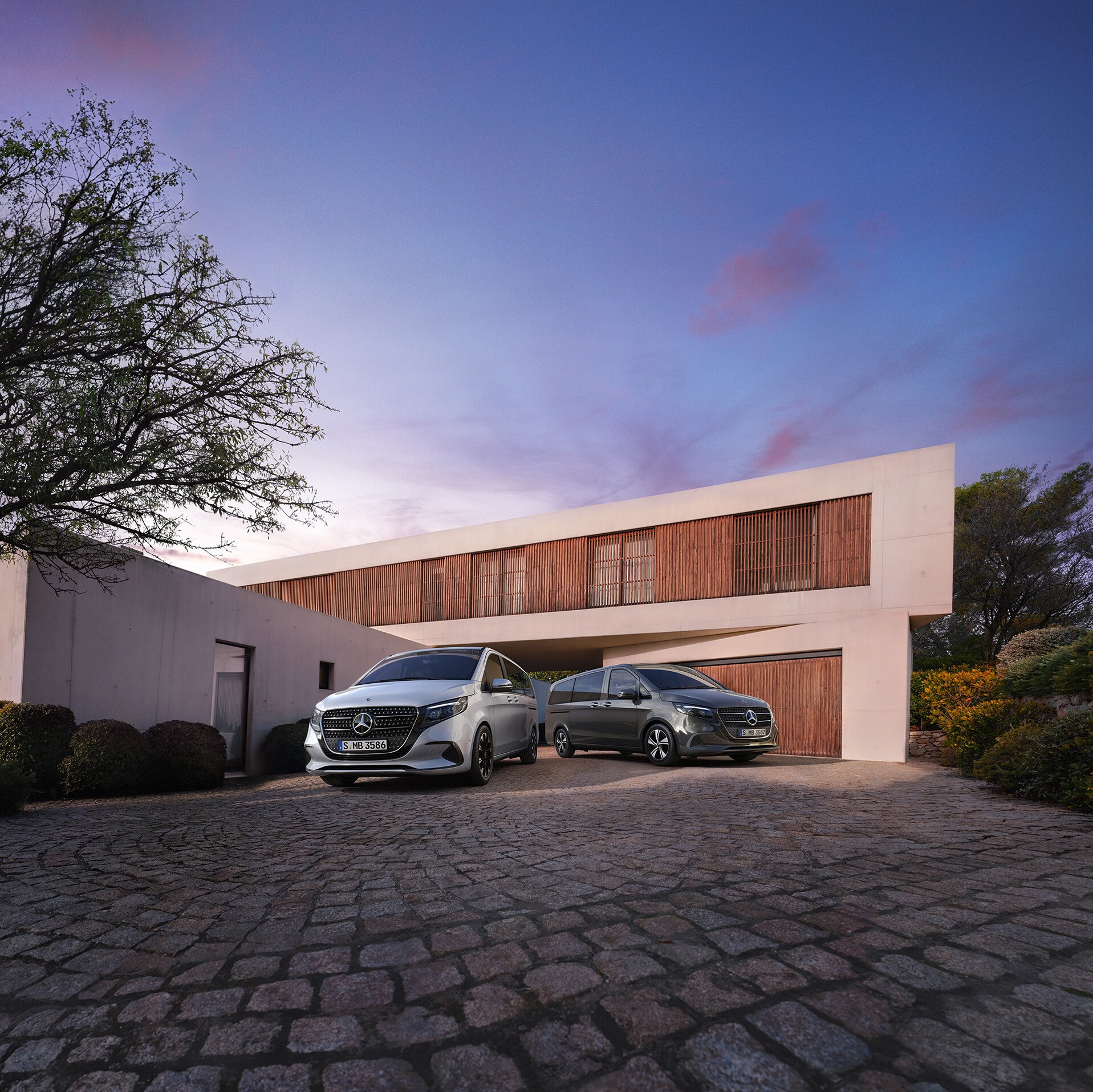 Zwei Mercedes-Benz V-Klasse parken vor einem modernen Luxushaus in der Abenddämmerung.