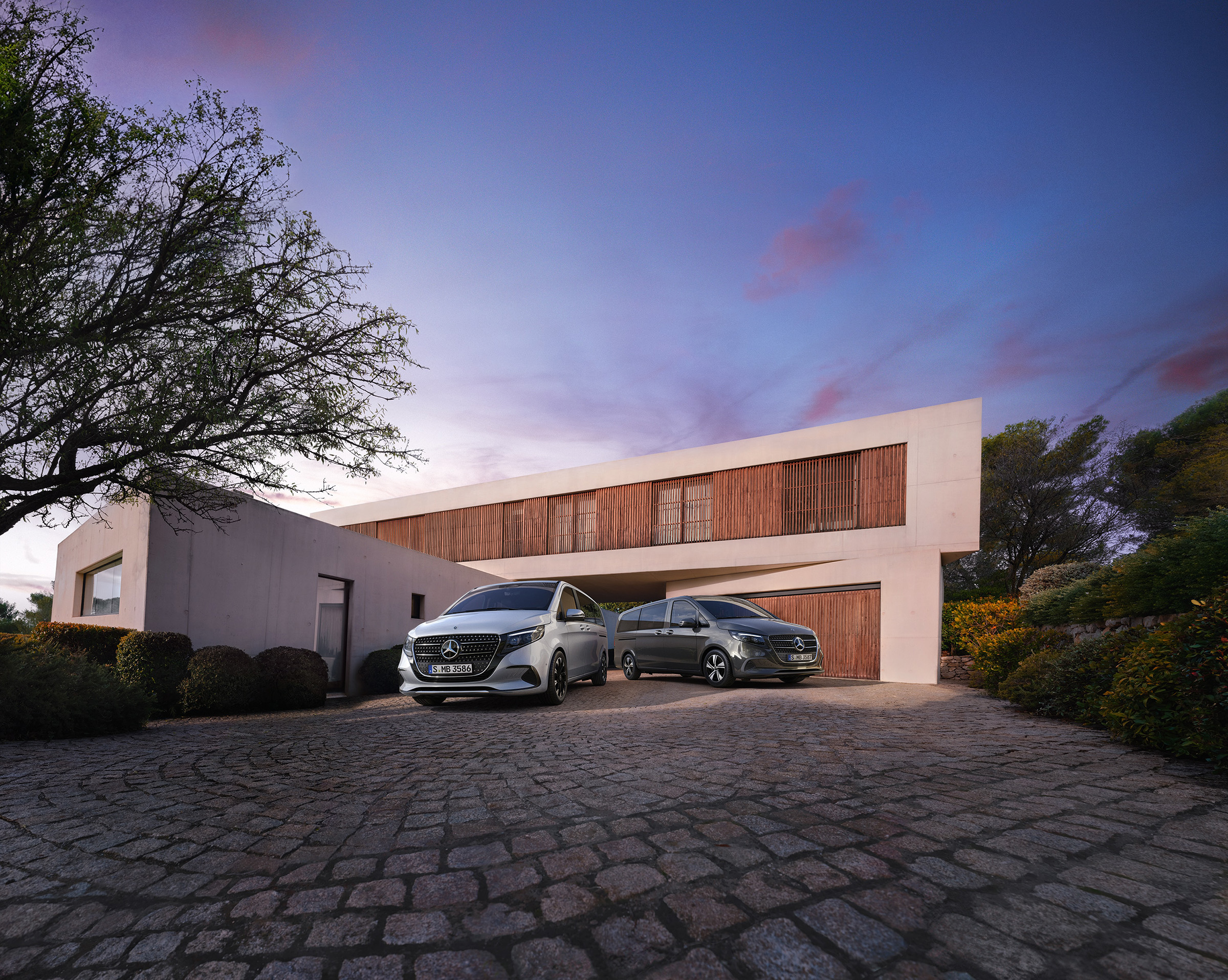 Van- und Camper-Modelle | Mercedes-Benz Vans. Zwei Mercedes-Benz V-Klasse parken vor einem modernen Luxushaus in der Abenddämmerung.