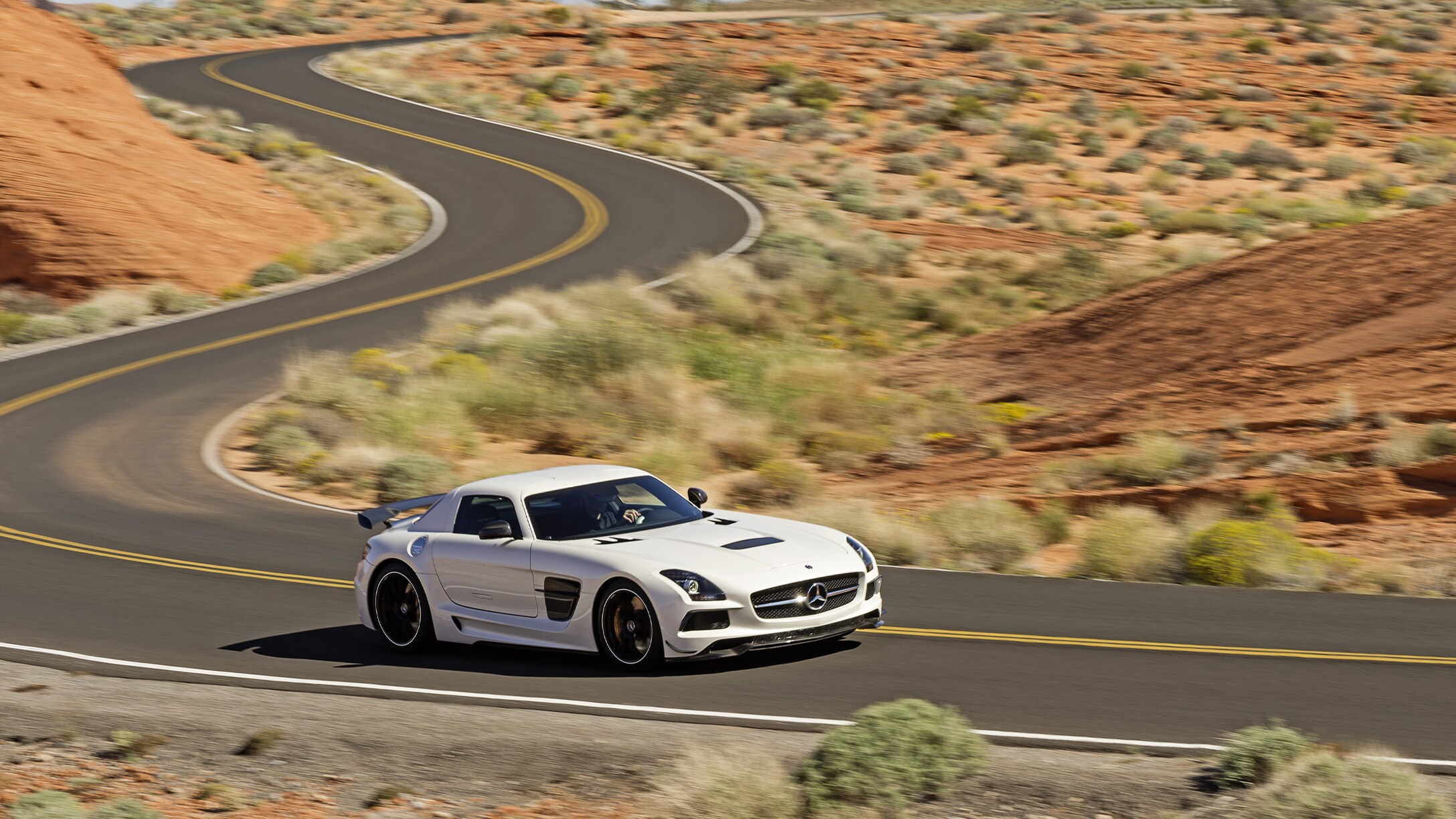 Ein weißer SLS AMG Black Series fährt auf einer leeren Straße durch eine wüstenähnliche Landschaft.