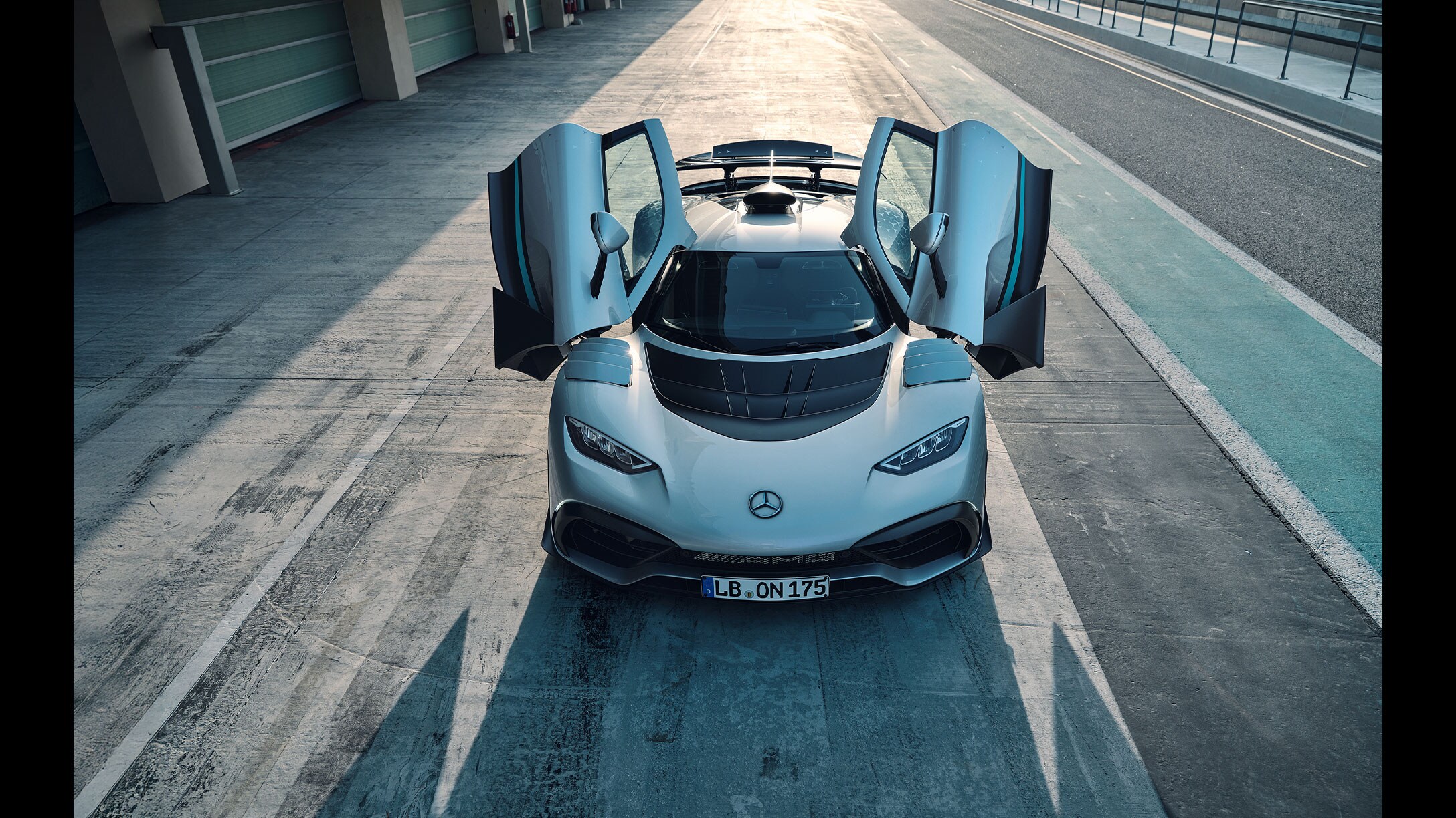 Das Bild zeigt eine Frontalansicht eines silbernen Mercedes-AMG ONE Hypercars mit geöffneten Flügeltüren. Das Fahrzeug steht in der Boxengasse einer Rennstrecke, mit den Boxengassen einer Rennstrecke.