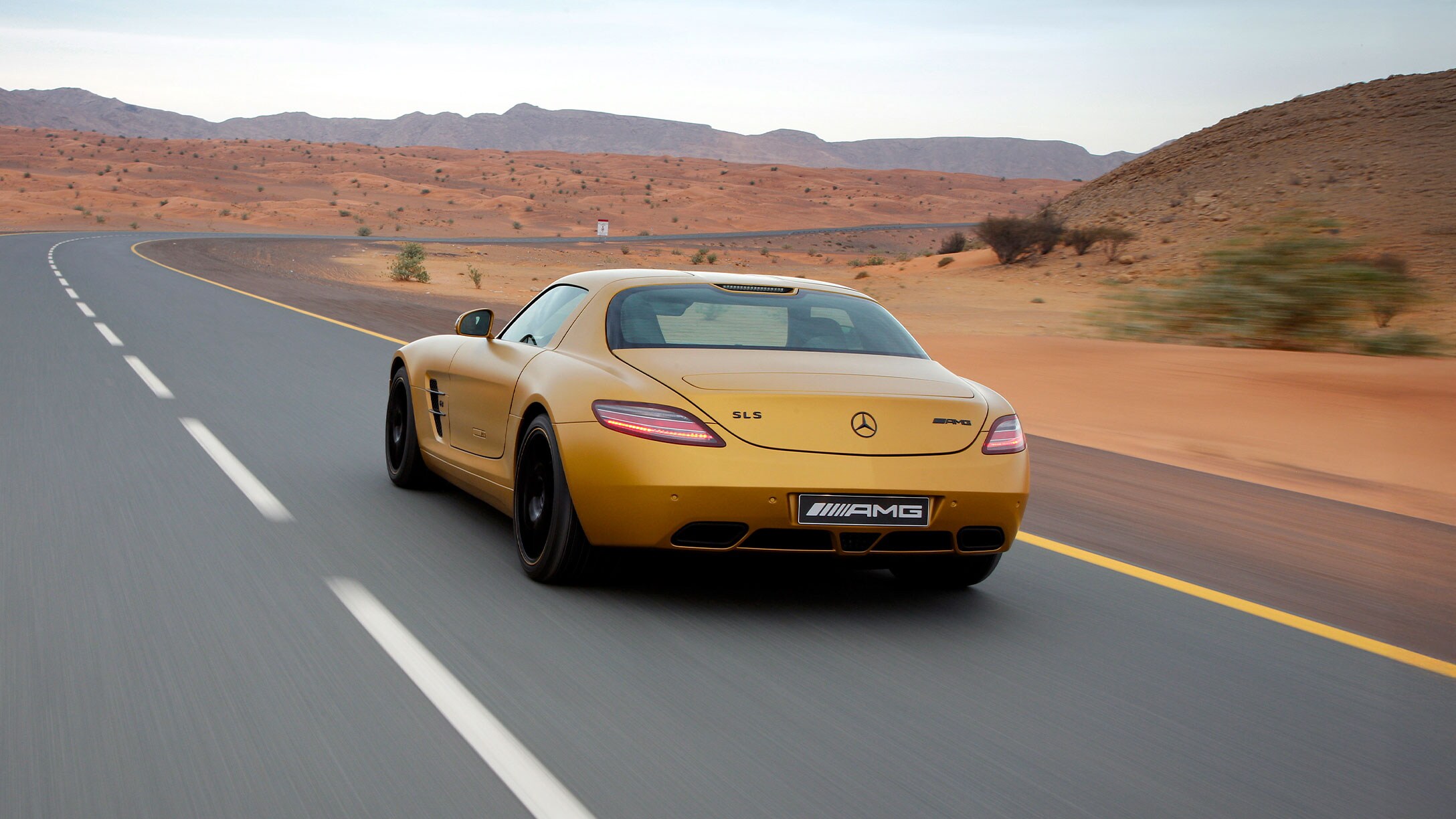 Ein orangener SLS AMG fährt auf einer leeren Straße durch eine wüstenähnliche Landschaft.