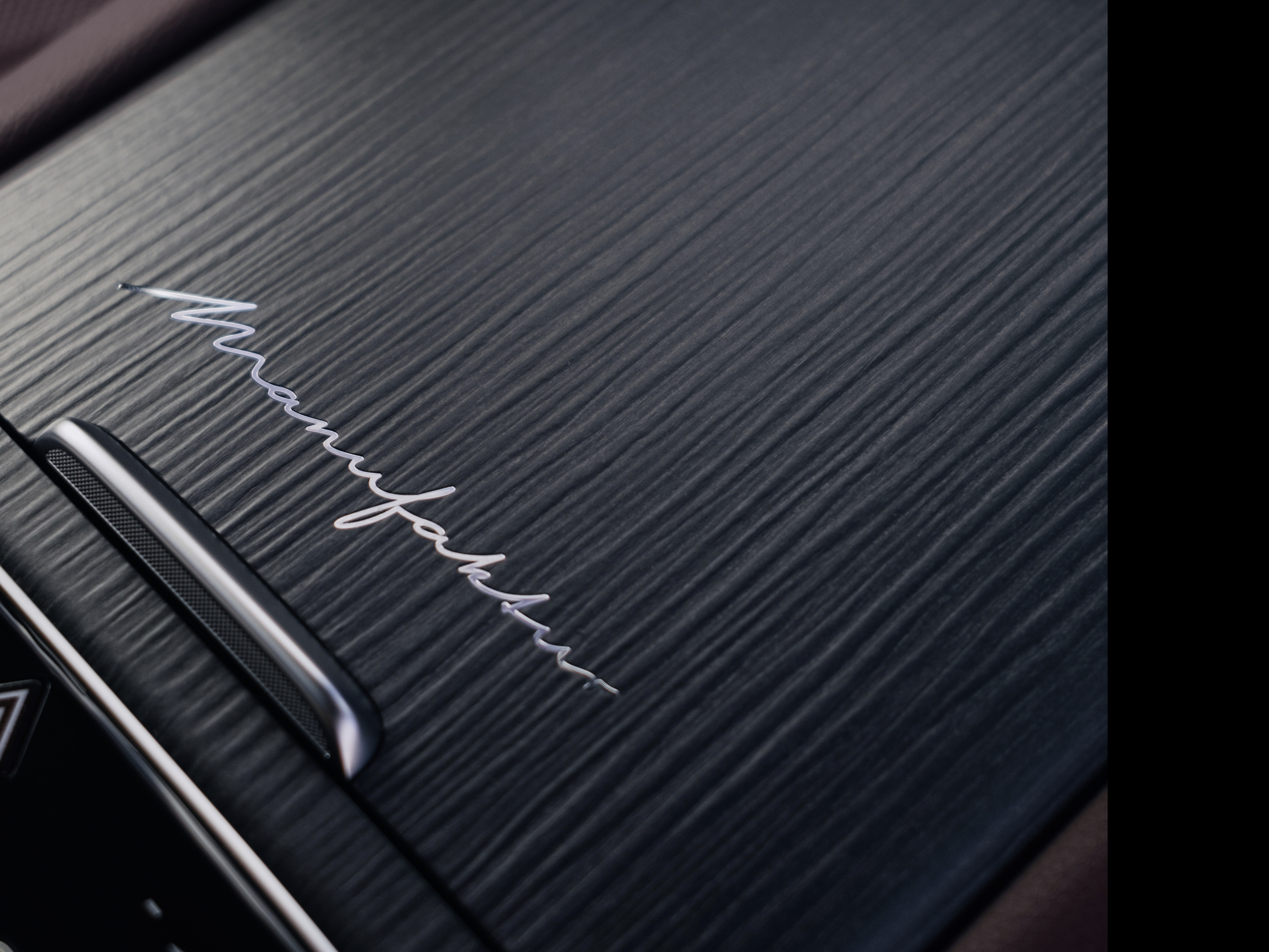 MANUFATUR | Die Marke | Mercedes-AMG Nahaufnahme einer dunkelgrauen, feingestreiften Holzoberfläche in einem Fahrzeuginnenraum mit dem silbernen Schriftzug „Manufaktur“ in Schreibschrift.