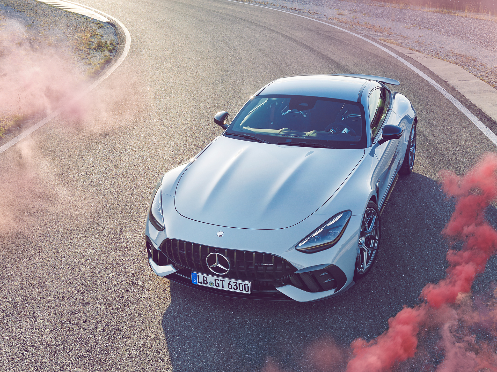 Motorsport | Die Marke | Mercedes-AMG Ein hellsilberner Mercedes-AMG GT Sportwagen auf einer Rennstrecke, aufgenommen aus der Vogelperspektive, umgeben von rosa Rauchschwaden.