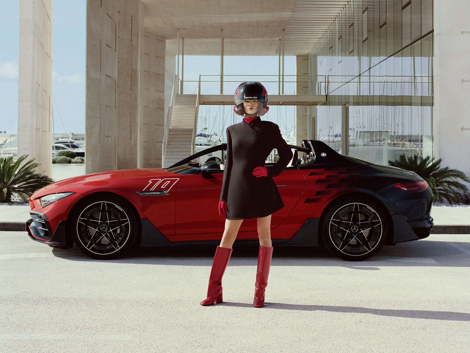 Ein offener Mercedes-AMG PureSpeed Roadster in Rot und Schwarz steht vor einem modernen Gebäude. Davor steht ein Model in einem futuristischen Outfit mit Integralhelm.