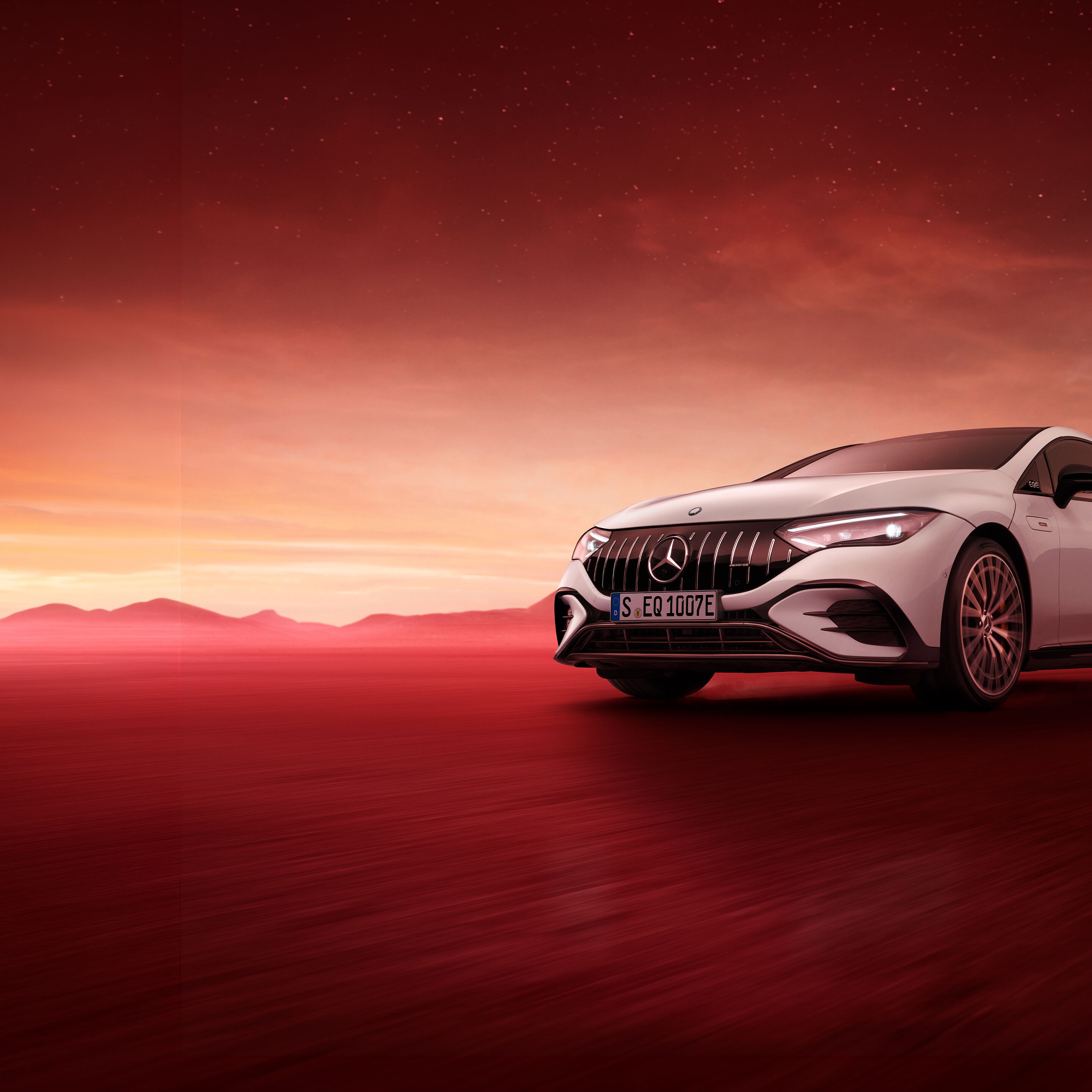 Frontalansicht eines weißen Mercedes-EQE, der auf einem roten Untergrund vor einer Bergkulisse und einem roten Sternenhimmel parkt.
