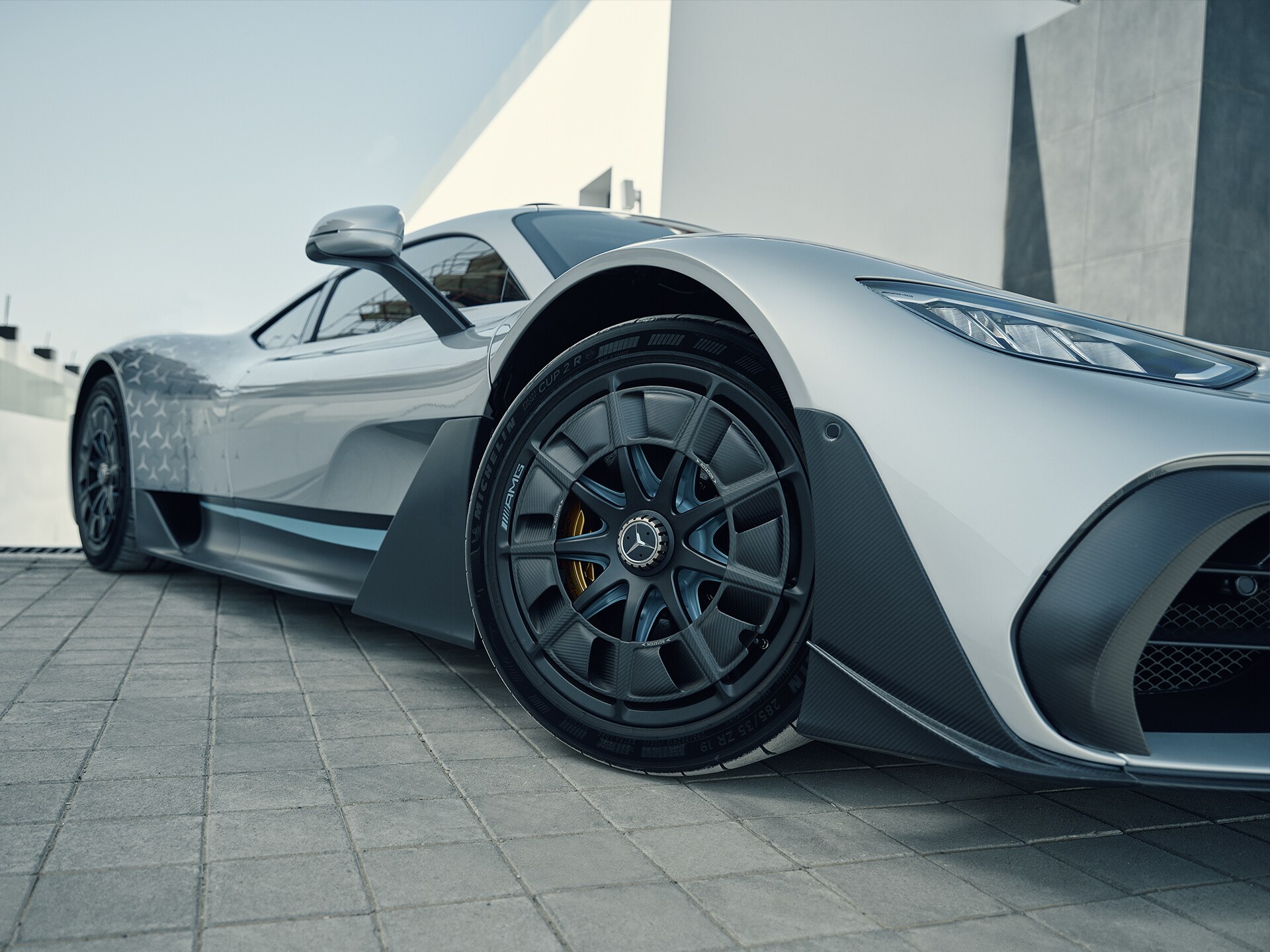 Nahaufnahme der vorderen Dreiviertelseite eines silbernen Mercedes-AMG ONE Hypercars, das auf einem gepflasterten Vorplatz vor einem weißen Gebäude parkt.