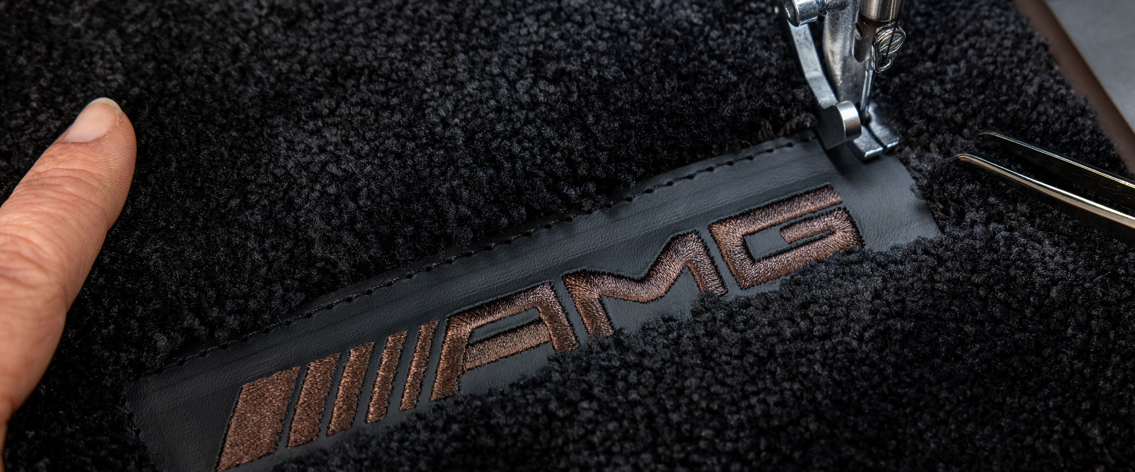 Ein brauner, gestickter Schriftzug "AMG", der auf einen Teppich von Mercedes-Benz genäht wird.