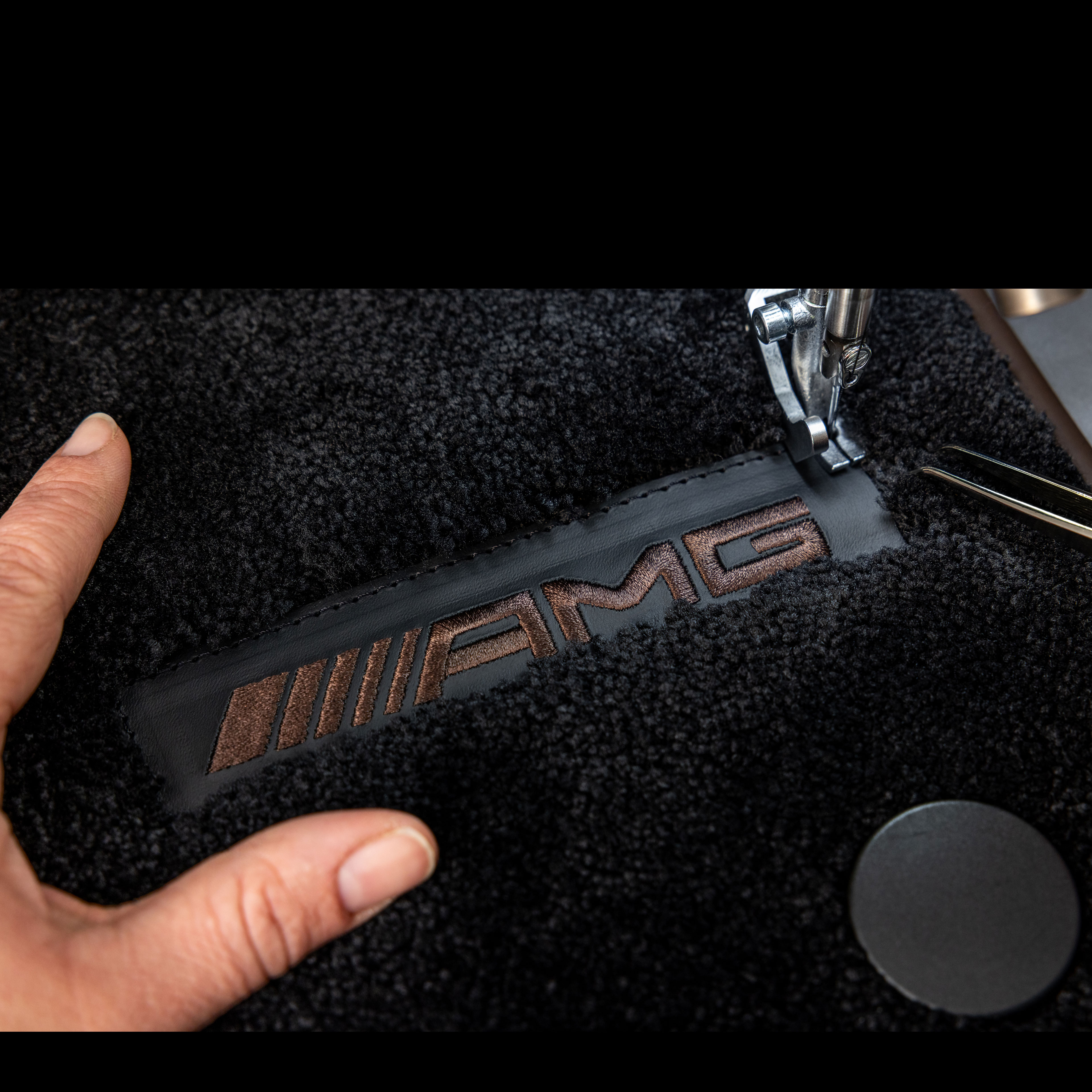 MANUFAKTUR | Mercedes-AMG Ein brauner, gestickter Schriftzug "AMG", der auf einen Teppich von Mercedes-Benz genäht wird.