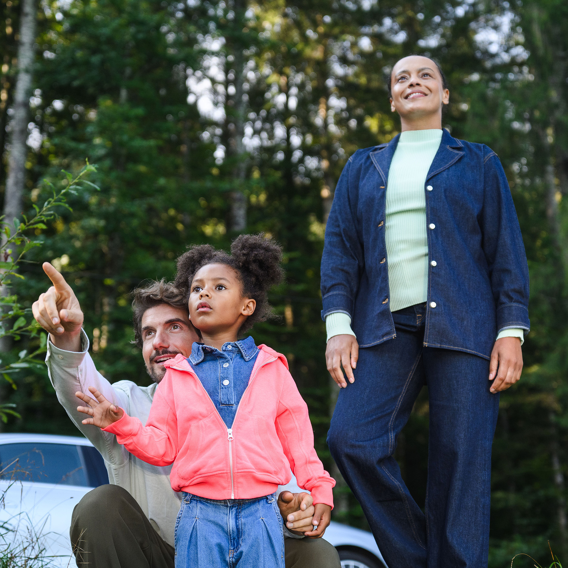 Buchungsplattform | Mercedes-Benz Rent Zwei Erwachsene und ein Kind stehen in einem Wald, im Hintergrund ist ein Mercedes-Benz Fahrzeug.