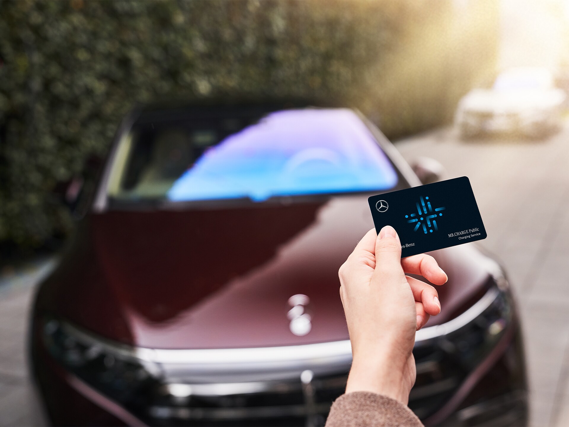 Eine MB.CHARGE Public Tankkarte in der Hand einer Person vor einem Mercedes-Benz.