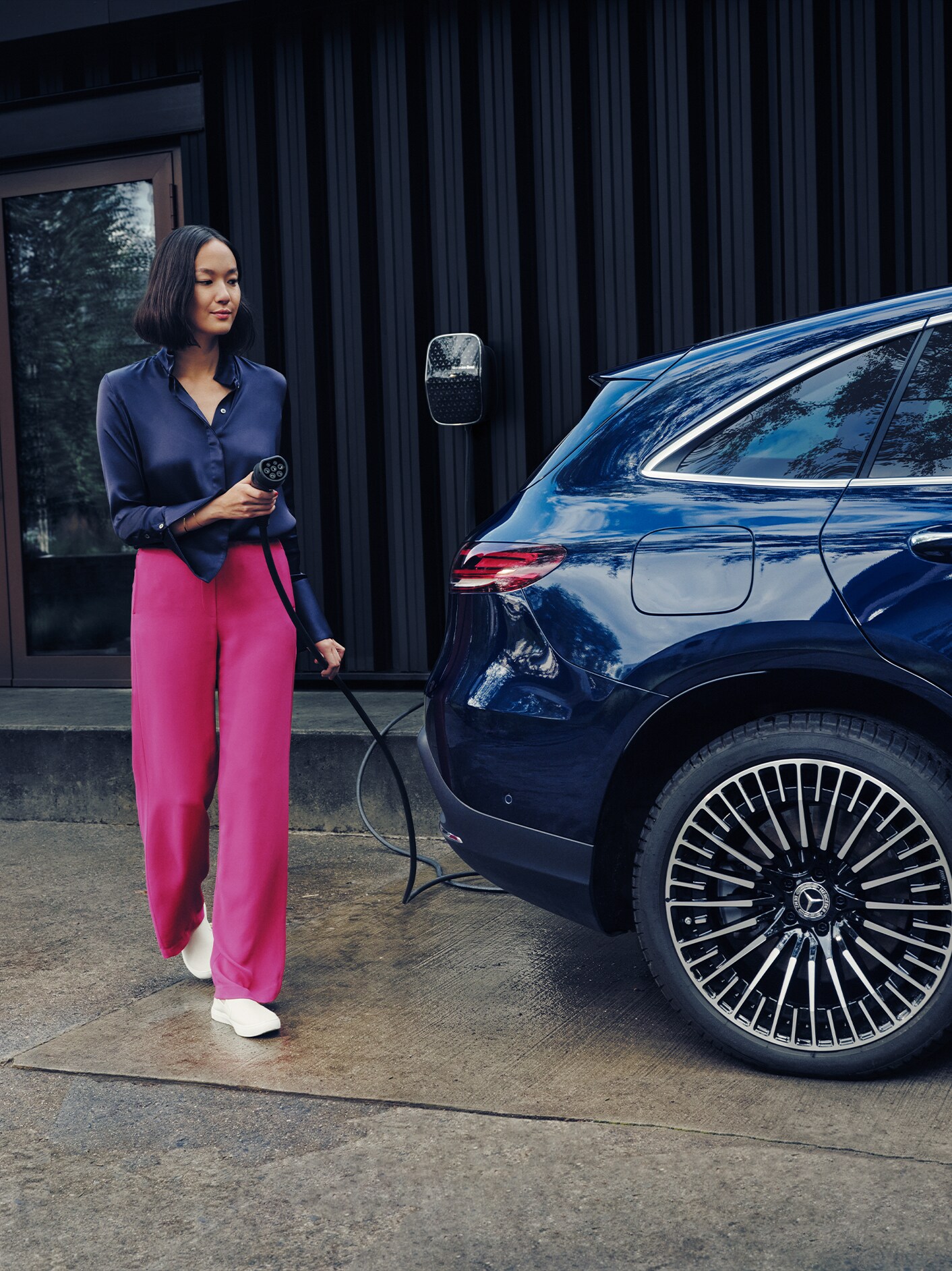 Eine Frau in pinker Hose lädt ein Mercedes-Benz Elektrofahrzeug mit einer Wallbox Ladestation auf.