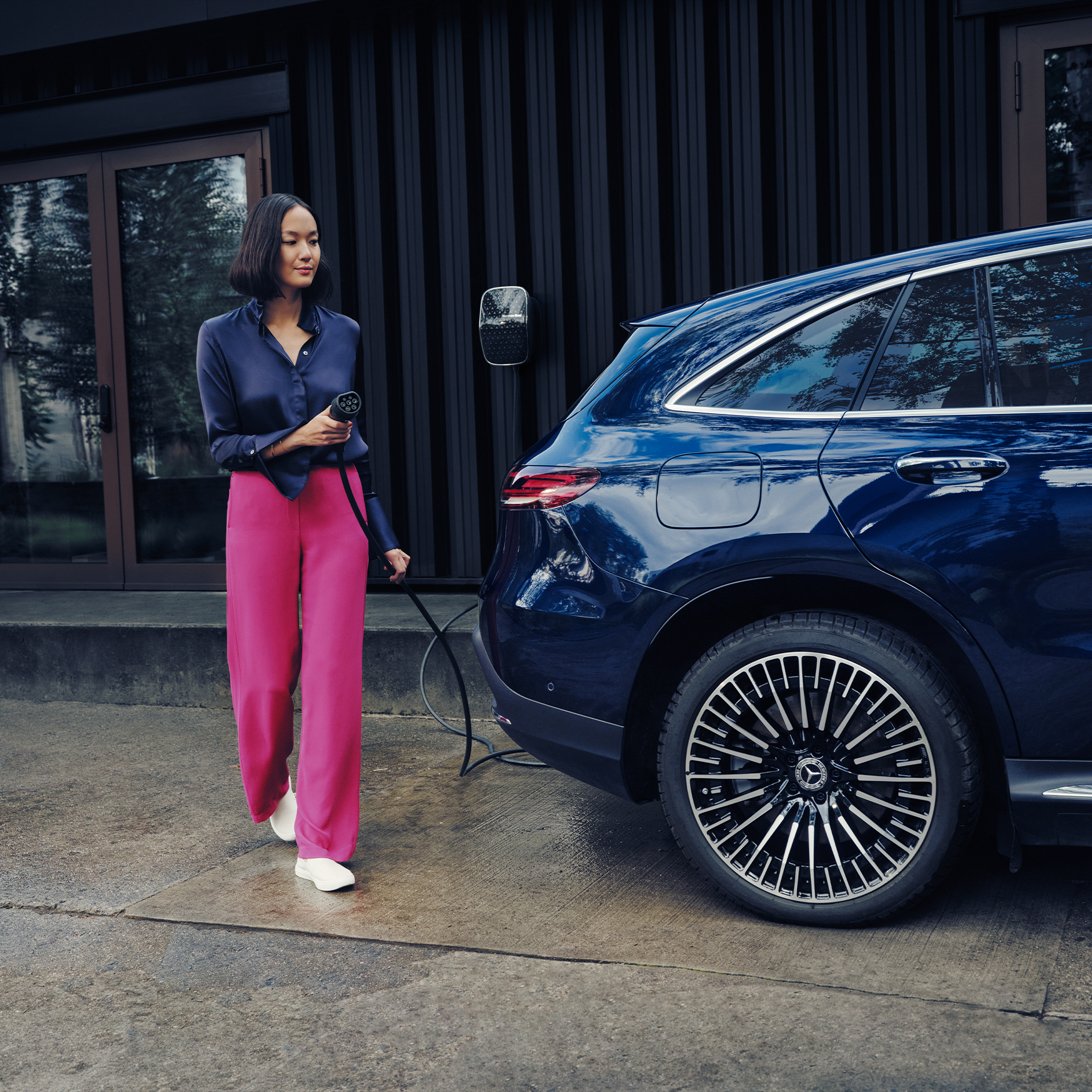 Ladelösungen für Unternehmen | Mercedes-Benz Eine Frau in pinker Hose lädt ein Mercedes-Benz Elektrofahrzeug mit einer Wallbox Ladestation auf.