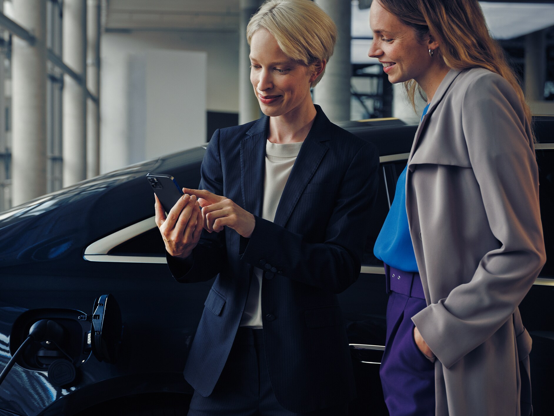 Zwei Frauen an einer modernen Arbeitsstätte betrachten gemeinsam ein Smartphone vor einer Mercedes-Benz EQS Limousine.