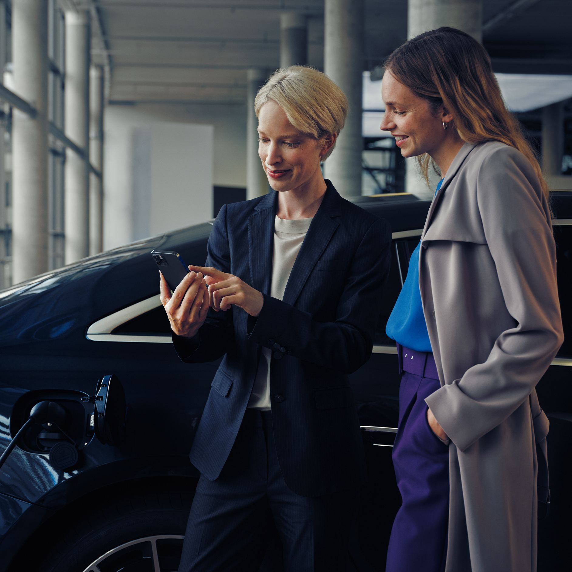 Ladelösungen für Unternehmen | Mercedes-Benz Zwei Frauen an einer modernen Arbeitsstätte betrachten gemeinsam ein Smartphone vor einer Mercedes-Benz EQS Limousine.