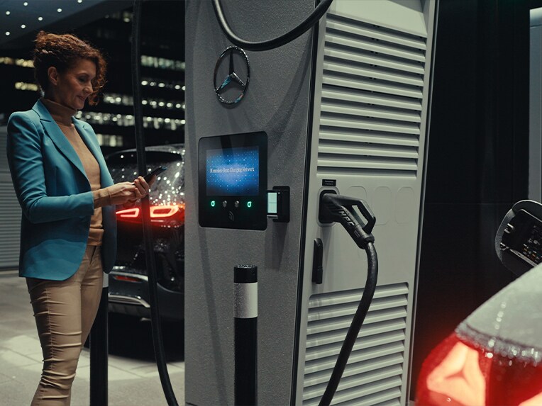 Eine Frau hält ihr Smartphone in der Hand und steht neben einer Ladestation.