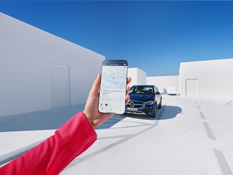 Eine Frau informiert sich per Mercedes-Benz App auf ihrem Smartphone und den Digitalen Extras zum Thema Navigation, wie das Wetter am Ankunftsort wird.
