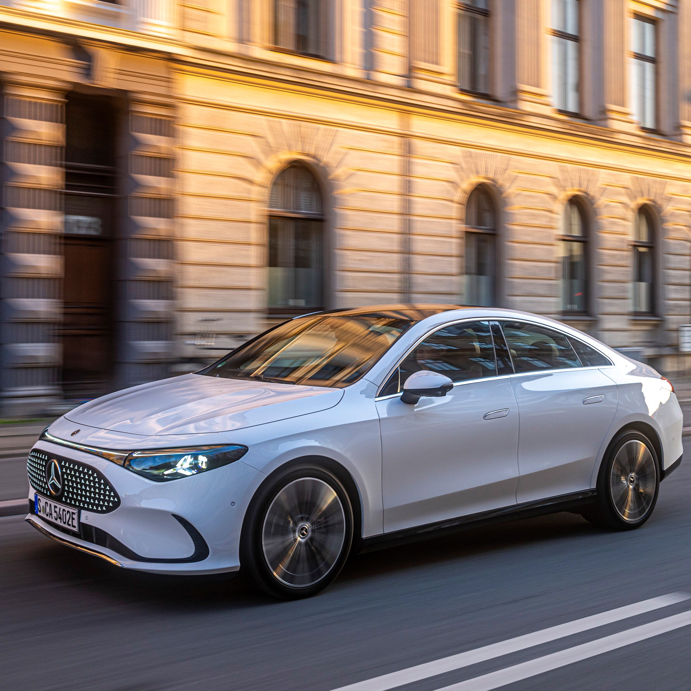 Ein heller Mercedes-Benz fährt an einem steinernen Gebäude bei untergehender Sonne entlang.