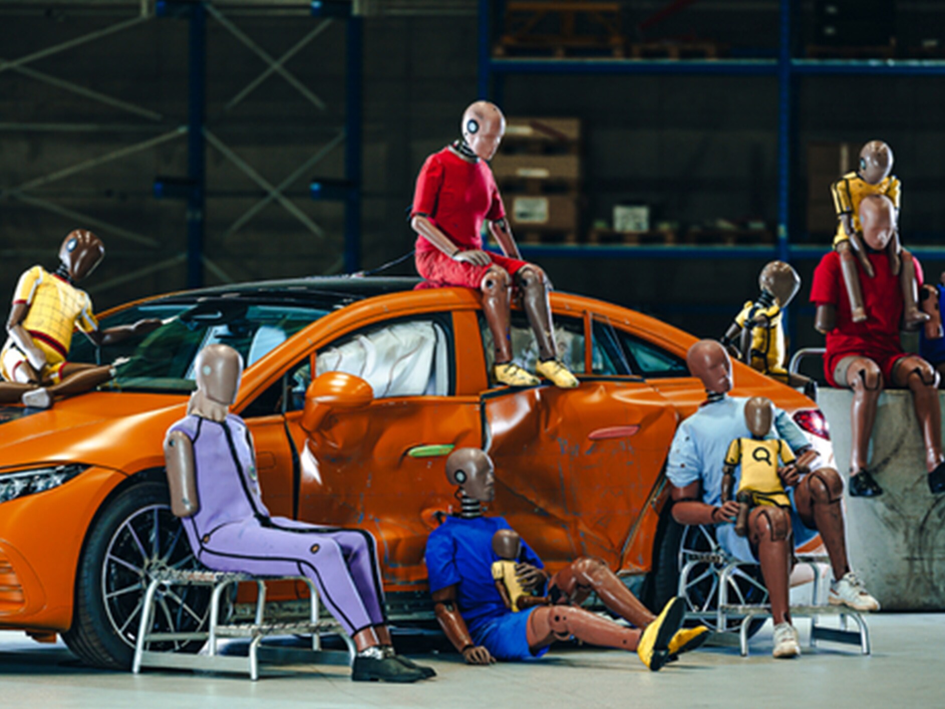 Mehrere Crashtest-Dummys sitzen auf und um ein orangenes Mercedes-Benz Auto mit zerbeulten Türen.
