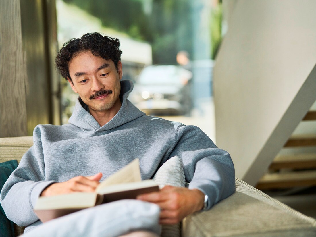 Ein Kunde sitzt in einem Haus und liest ein Buch. Durch ein Fenster hinter ihm ist ein Auto zu sehen, das von einem Spezialisten gewartet wird.