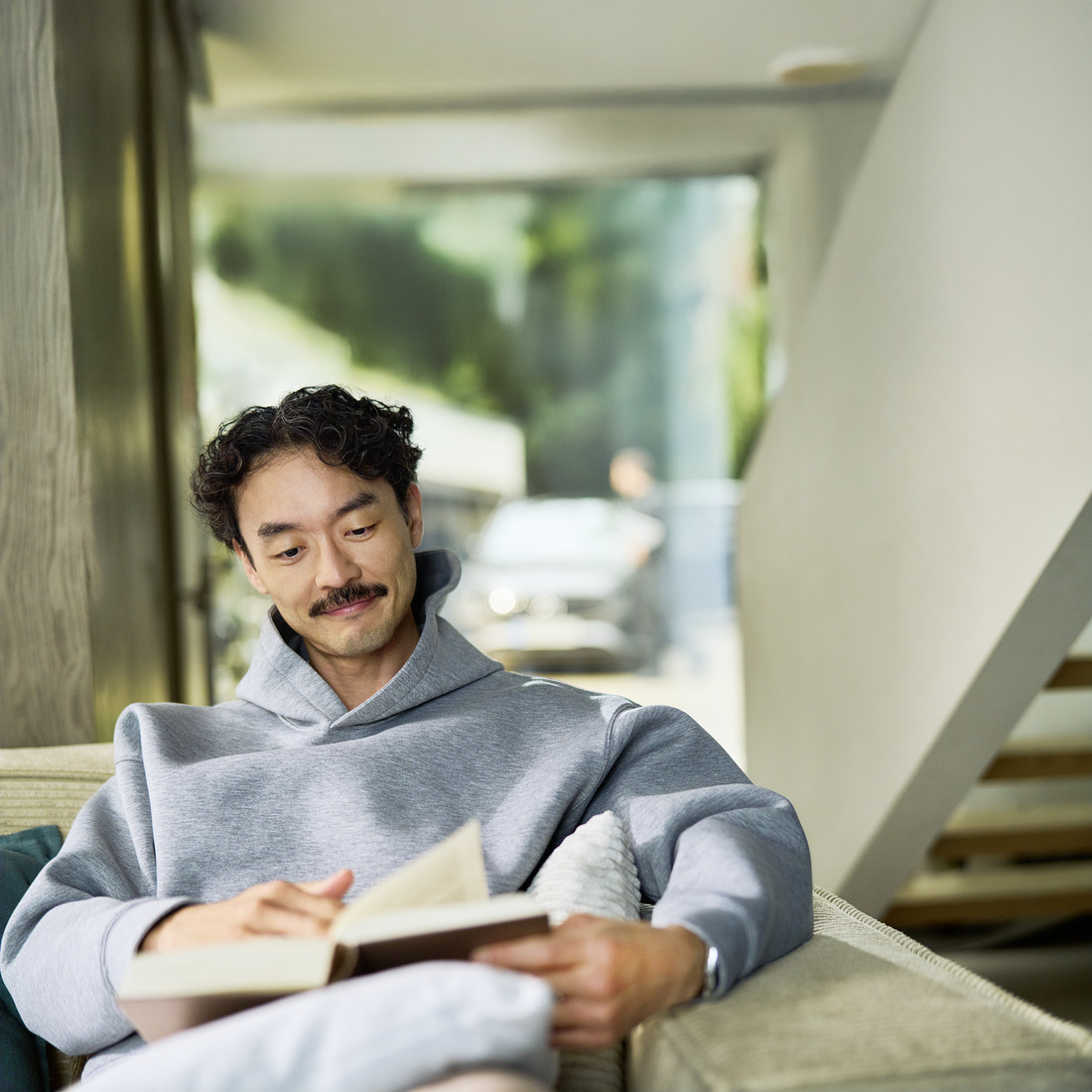 Entspannter Kunde, der Mobile Service zu Hause genießt Ein Kunde sitzt in einem Haus und liest ein Buch. Durch ein Fenster hinter ihm ist ein Auto zu sehen, das von einem Spezialisten gewartet wird.