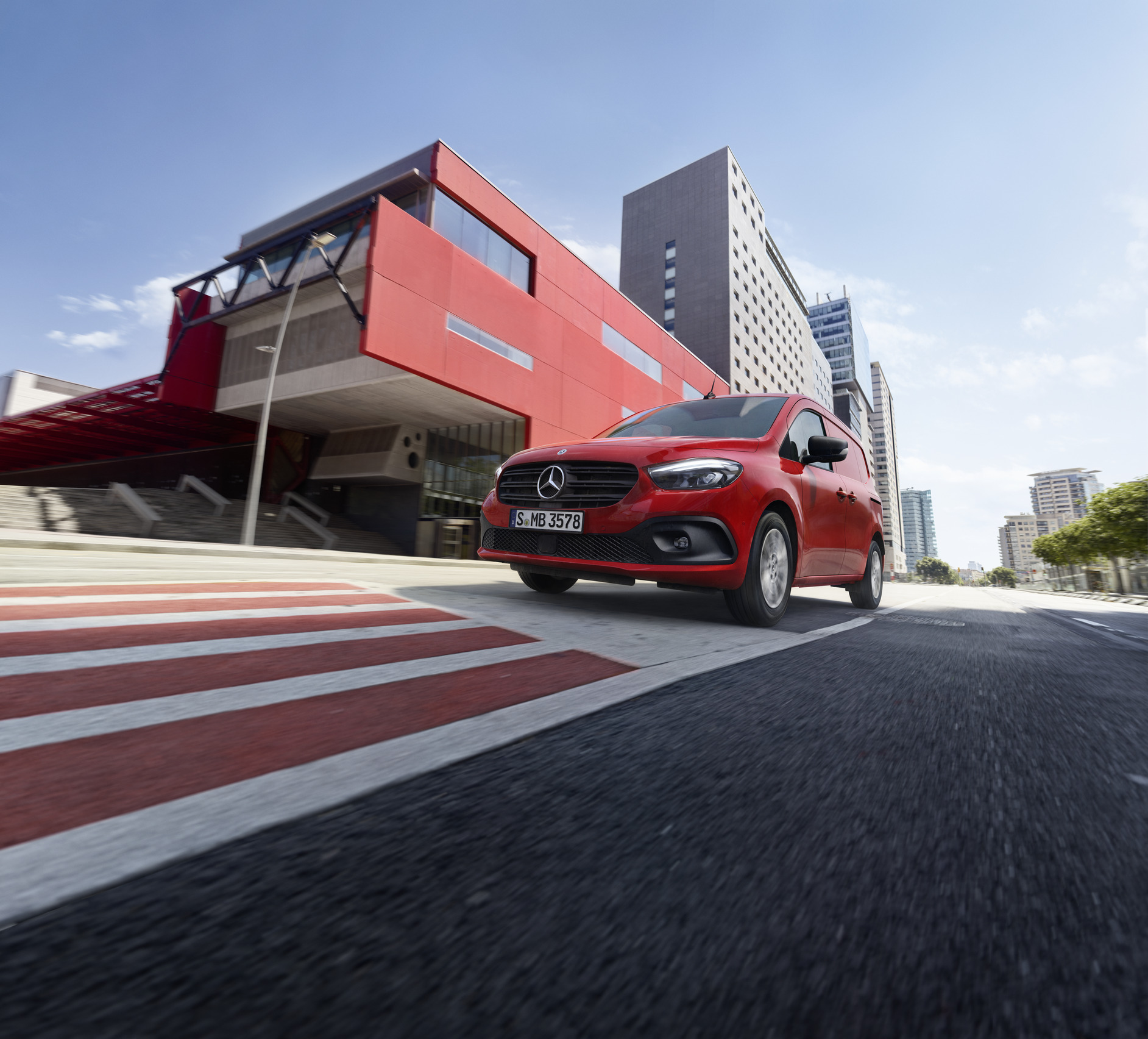 Assistenz- & Sicherheitssysteme | Mercedes-Benz Transporter. Ein roter Mercedes-Benz Citan Kastenwagen ist auf der Straße unterwegs neben einem roten Gebäude.