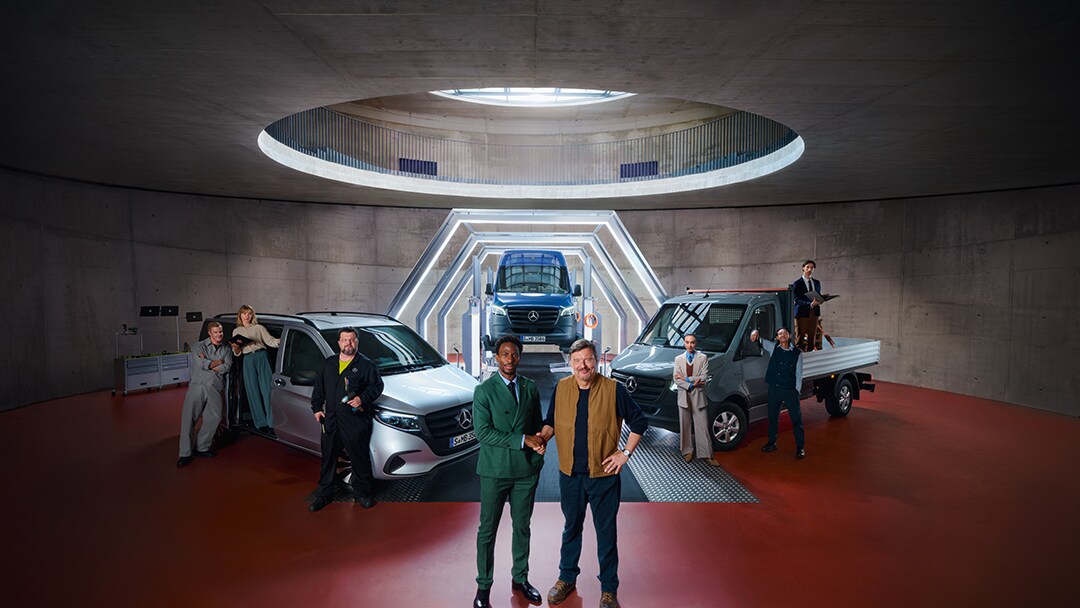 Eine Gruppe Menschen steht im einem Raum vor Mercedes-Benz Transportern. Von oben scheint Licht.