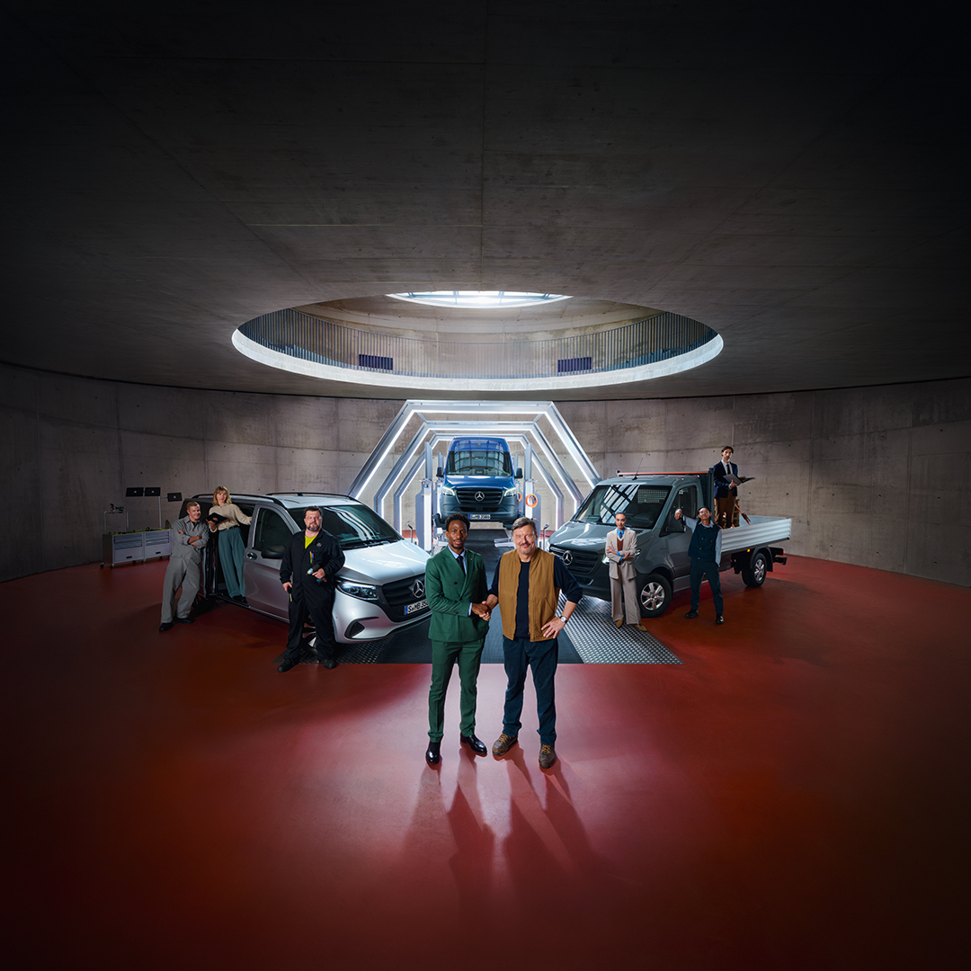 Eine Gruppe Menschen steht im einem Raum vor Mercedes-Benz Transportern. Von oben scheint Licht.