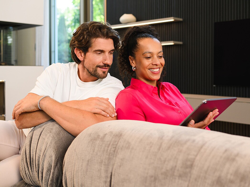 Eine Frau mit pinker Bluse und ein Mann mit weißem T-Shirt sitzen auf der Couch und schauen auf ein Tablet.