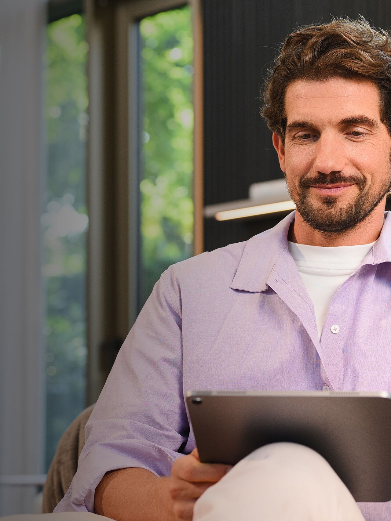 Ein lächelnder Mann mit fliederfarbenem Hemd sitzen auf der Couchund schaut auf ein Tablet.