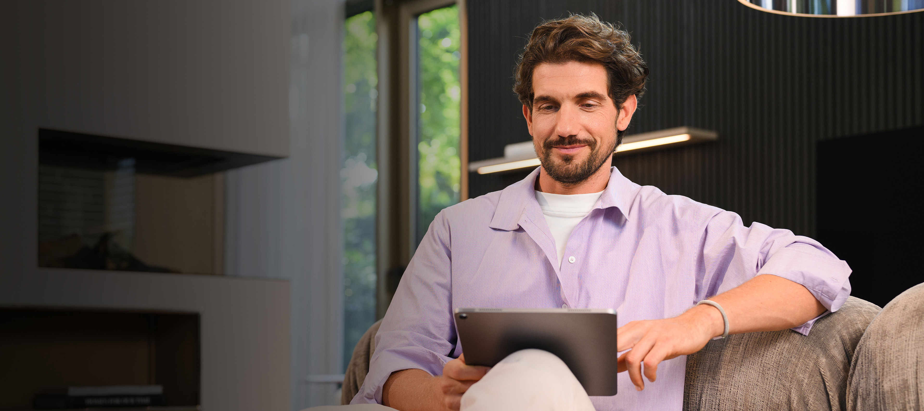 Mercedes-Benz finance | Mercedes-Benz. Ein lächelnder Mann mit fliederfarbenem Hemd sitzen auf der Couchund schaut auf ein Tablet.