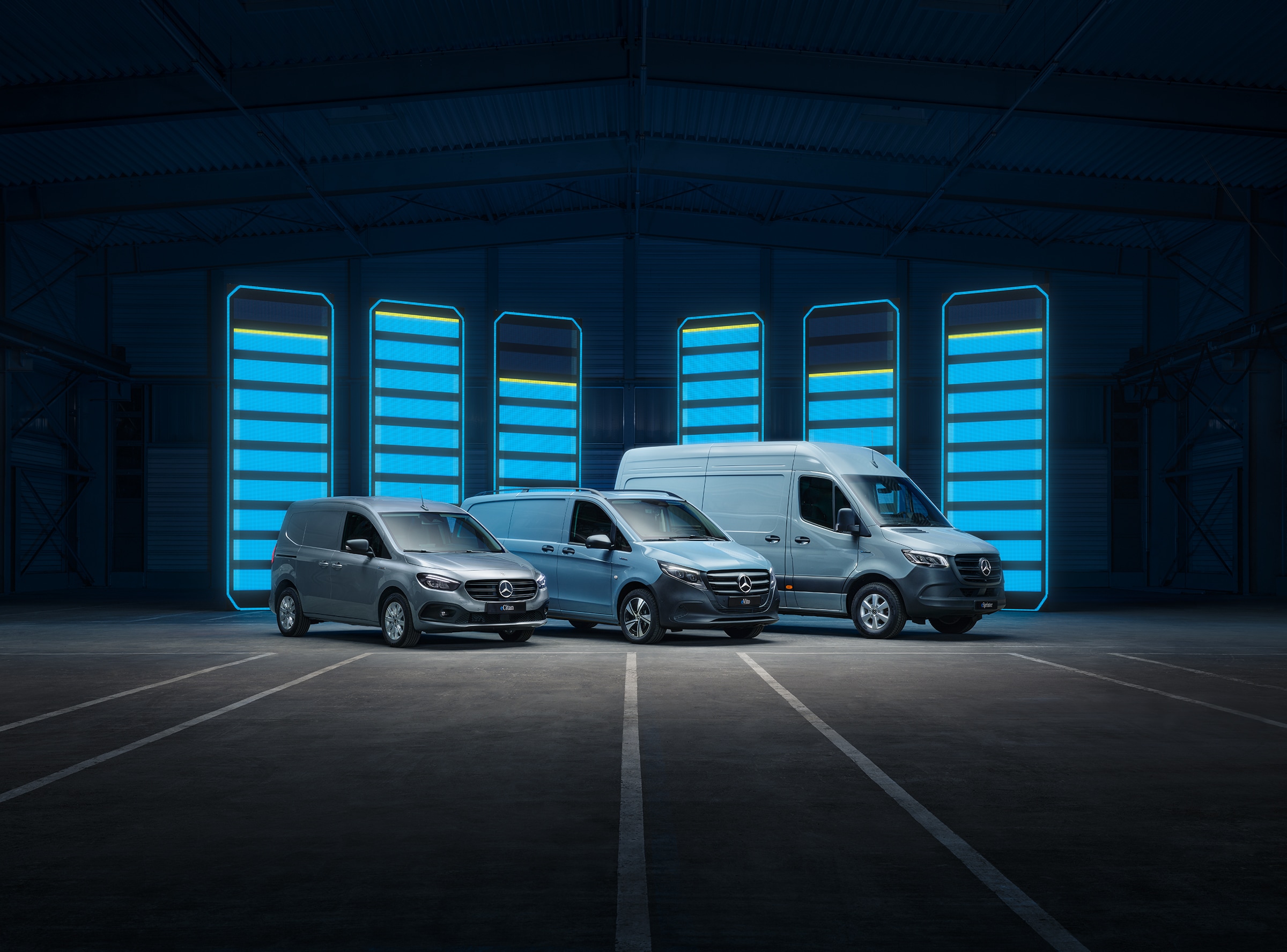 Geschichte | Mercedes-Benz Transporter. Die Elektrotransporter-Modellpalette von Mercedes-Benz, ausgestellt in einer Lagerhalle mit beleuchteten Ladestationen.