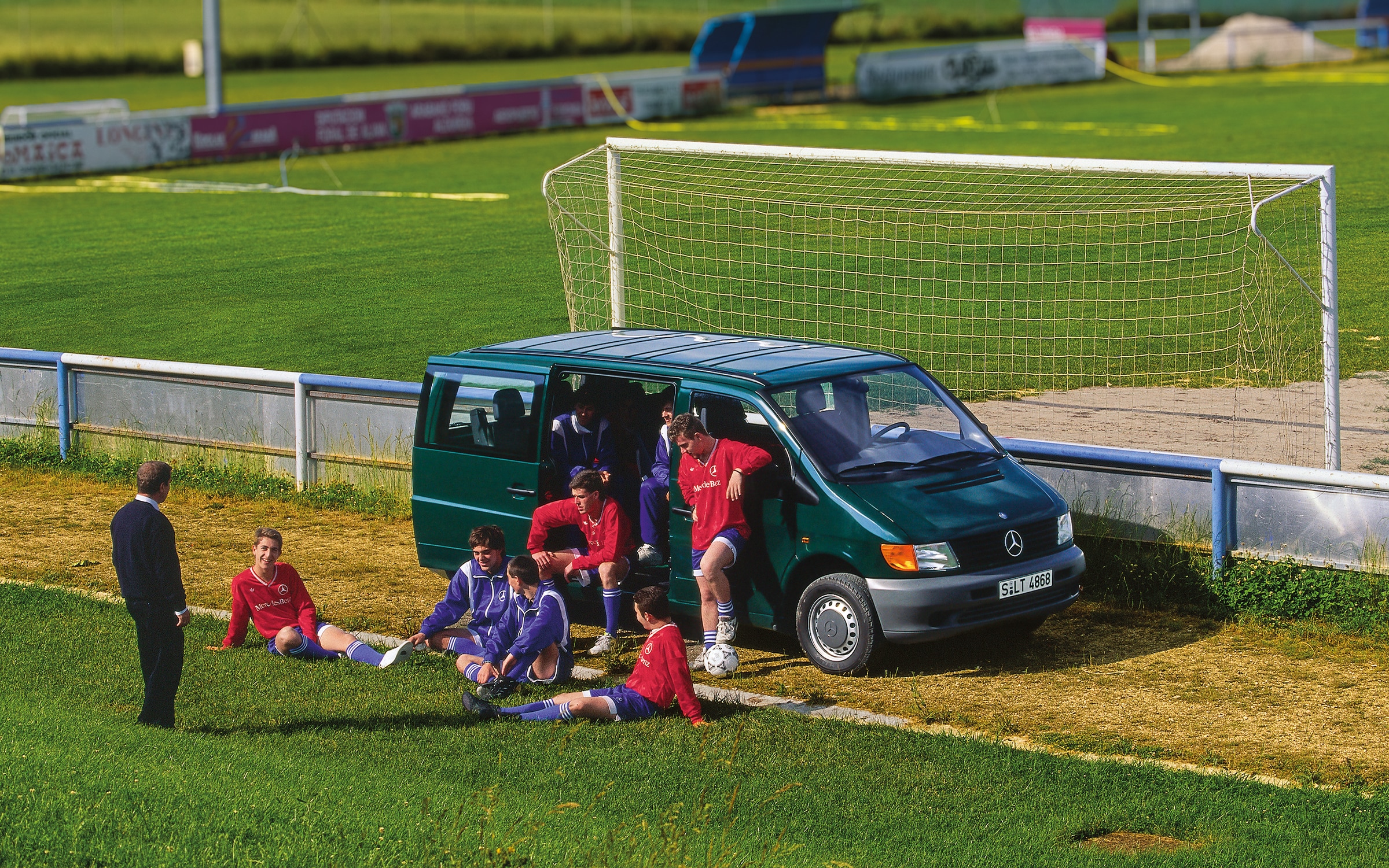 Geschichte | Mercedes-Benz Transporter. Ein Vito der ersten Generation, der neben einem Fußballplatz geparkt ist, mit Spielern der Mannschaft in der Nähe.
