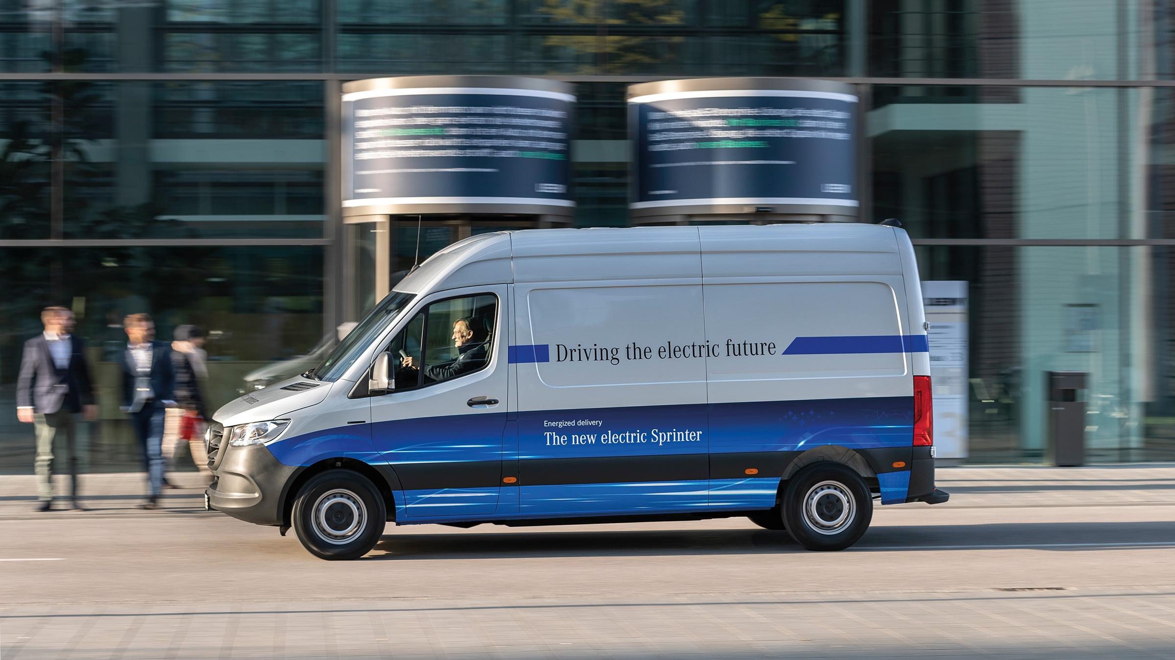 Geschichte | Mercedes-Benz Transporter. Ein Mercedes-Benz eSprinter Kastenwagen fährt durch eine moderne Stadtstraße.