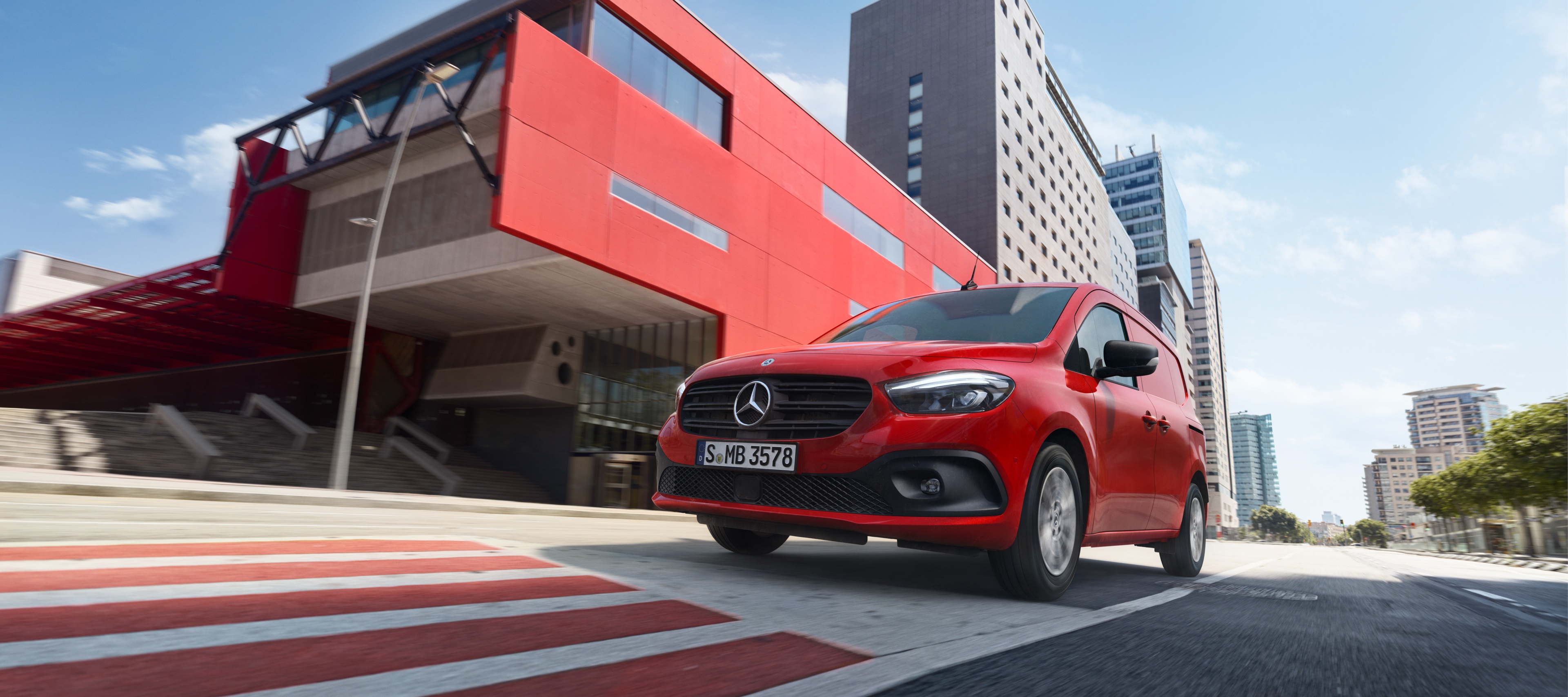 Citan Kastenwagen | Mercedes-Benz Transporter. Ein roter Citan Kastenwagen fährt auf einer Straße neben einem roten Gebäude.