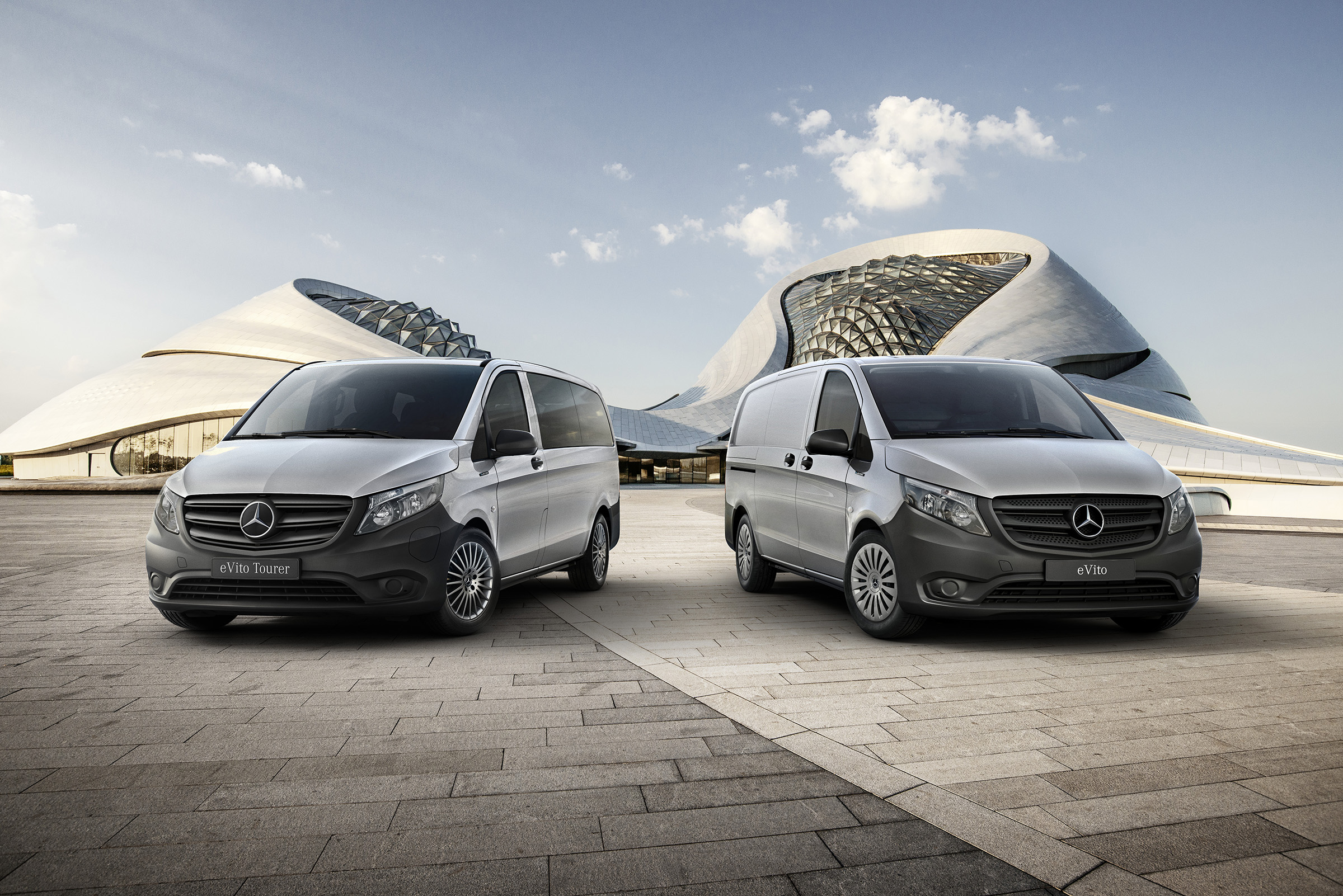 eVito Kastenwagen und Tourer | Alle Vito-Modelle | Mercedes-Benz Ein grauer eVito Kastenwagen und ein grauer Tourer vor einem futuristischen Gebäude auf einem gepflasterten Weg.