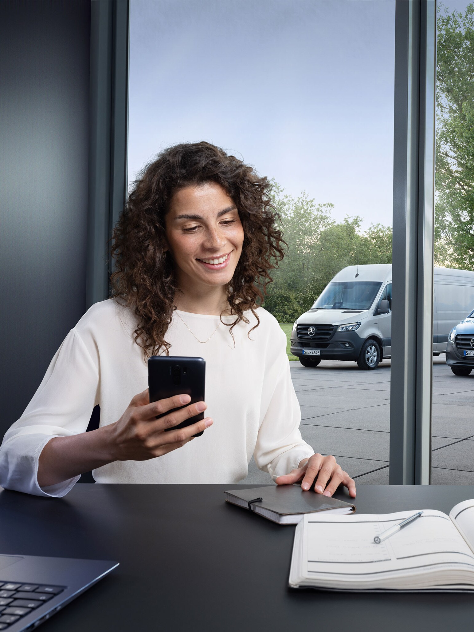 Frau am Schreibtisch mit Smartphone, im Hintergrund Vans – kompetente Betreuung im Business Care-Bereich.
