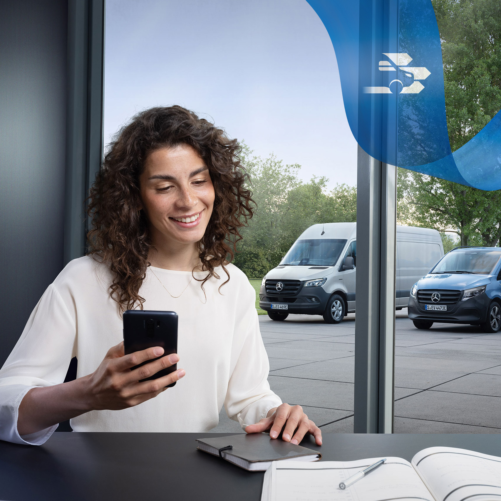 Customer Assistance Center | Mercedes-Benz Transporter. Frau am Schreibtisch mit Smartphone, im Hintergrund Vans – Kundenbetreuung im Business Care-Bereich.