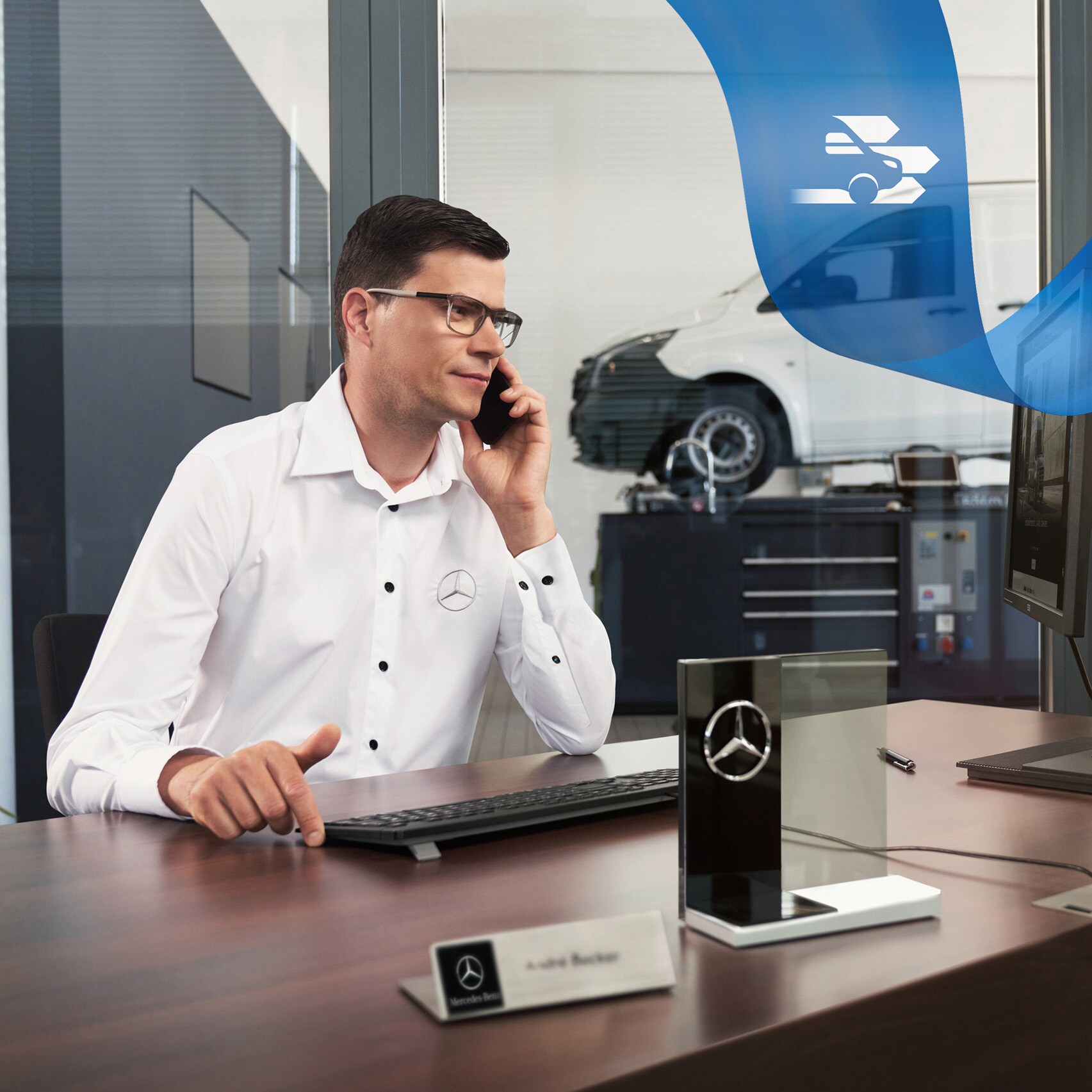 Mitarbeiter am Schreibtisch telefoniert für kompetente Betreuung im Care-Bereich, Van in der Werkstatt im Hintergrund.