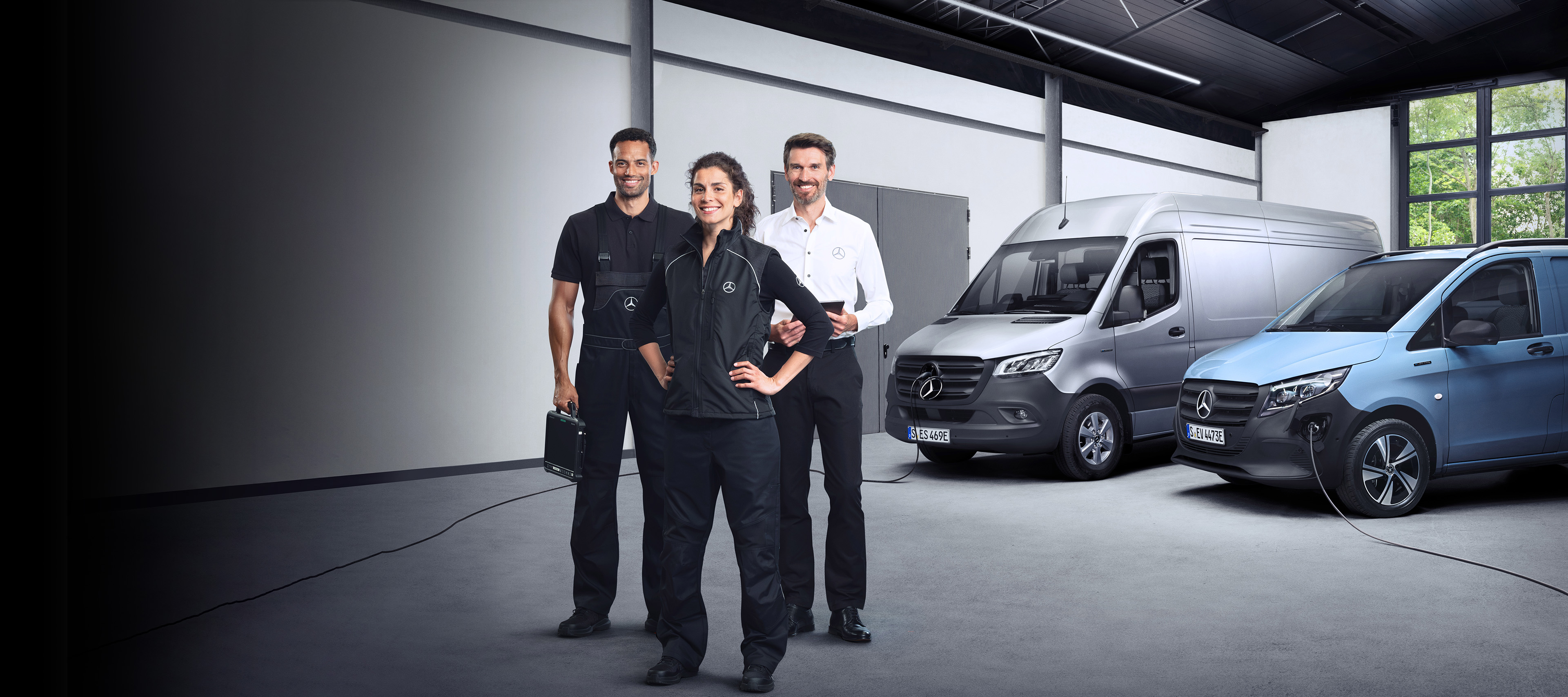 Elektrotransporter | Mercedes-Benz Drei Mercedes-Benz Elektrotransporter in einem dunklen Raum vor einer blauen Leuchttafel mit Gesichter von Arbeitern.